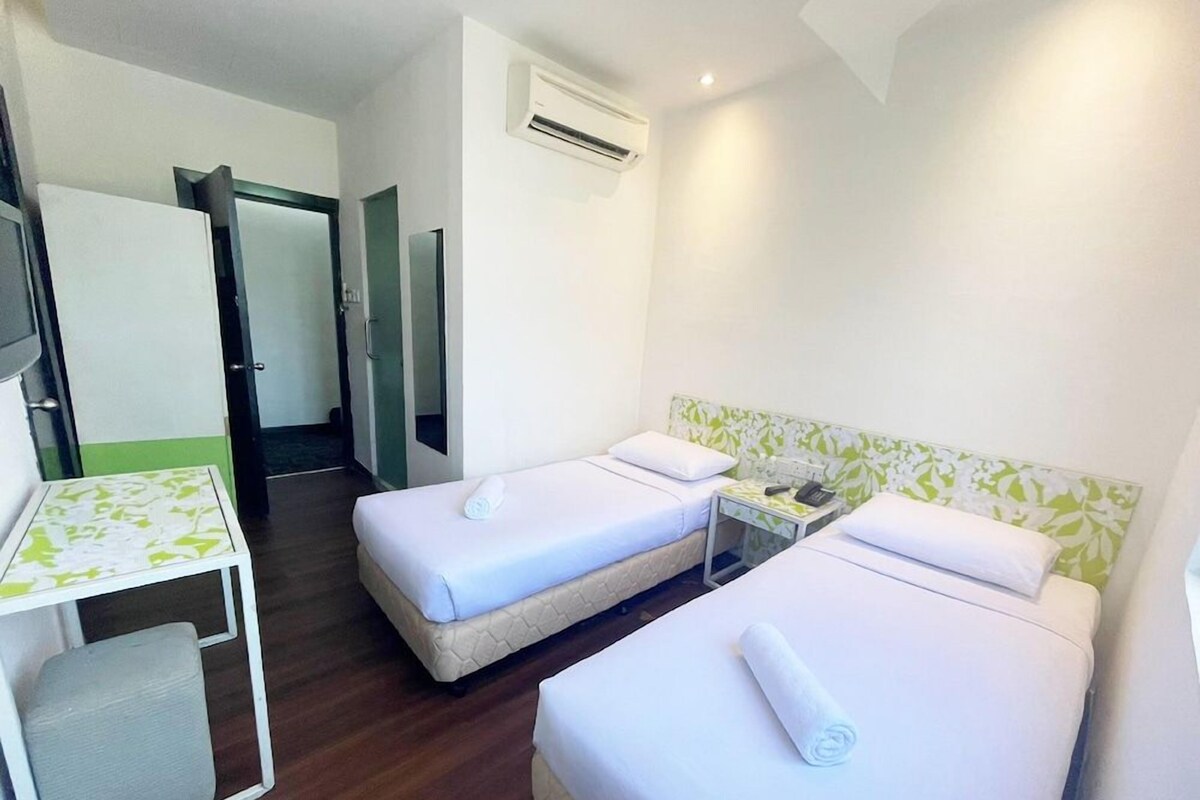 A hotel room with two single beds, a small table with a chair, an air conditioning unit, and a door leading to another room.