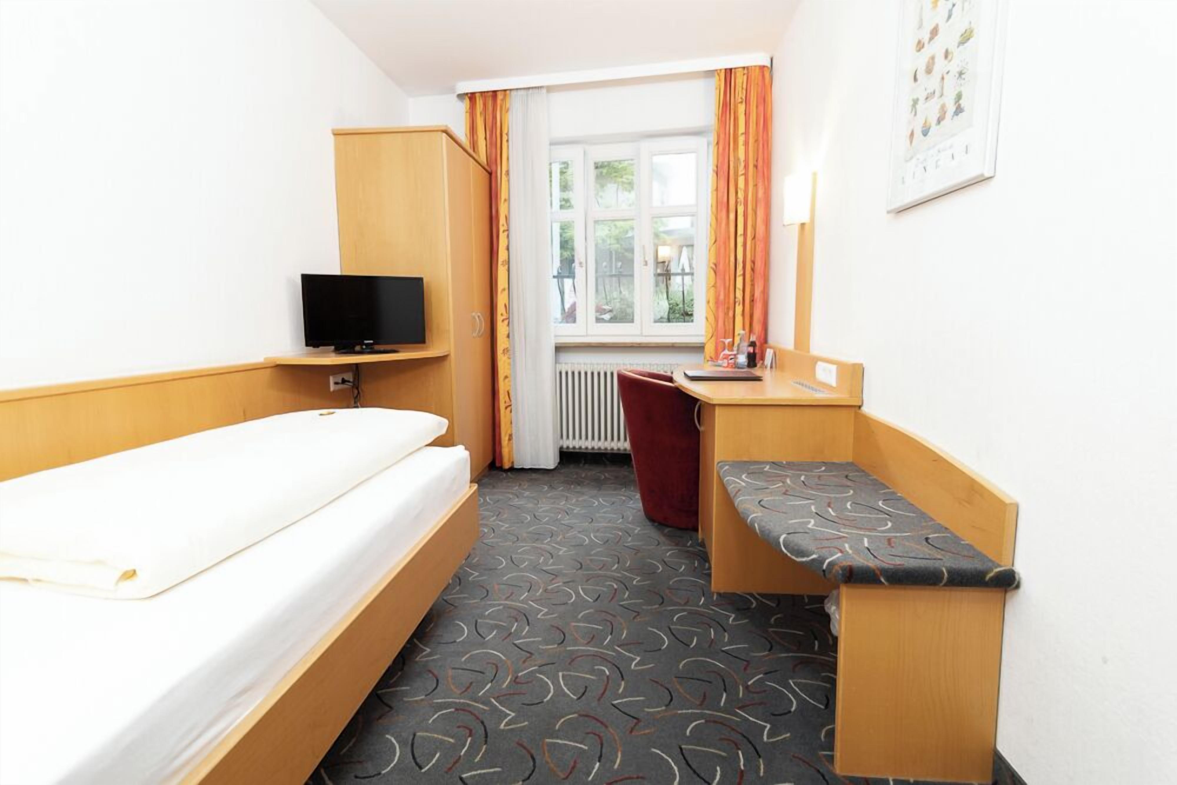 Une chambre d’hôtel de taille réduite, comprenant un lit simple, un bureau avec une chaise, une télévision et une fenêtre avec des rideaux.