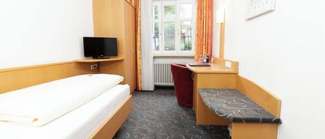 Une chambre d’hôtel de taille réduite, comprenant un lit simple, un bureau avec une chaise, une télévision et une fenêtre avec des rideaux.