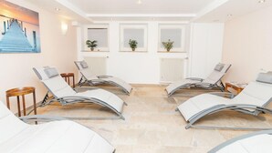 A sunlit room with four reclining chairs, a painting of a pier, and three windows with plants. - Hotel Garni Brugger (Lindau)
