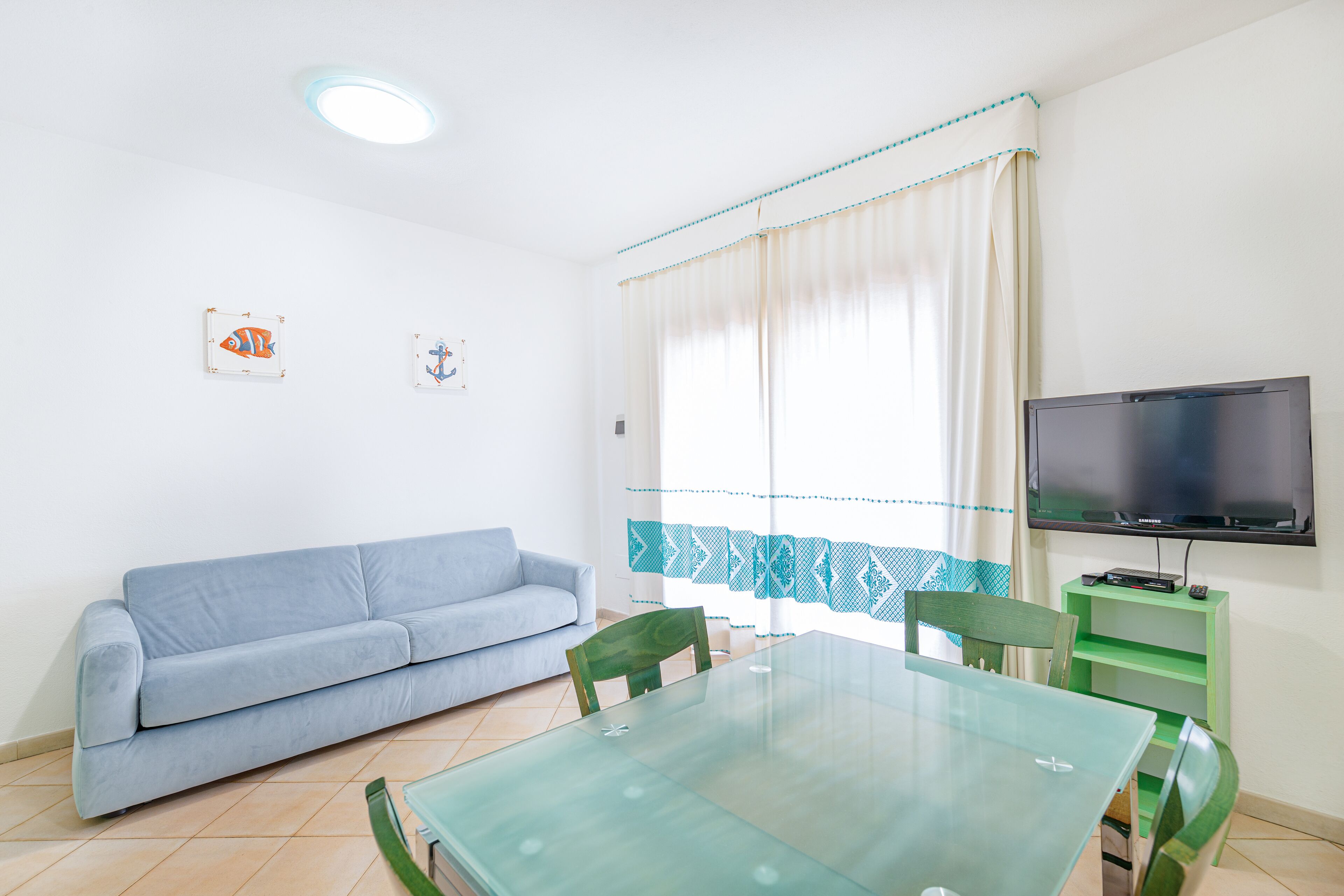 Un salon avec une table en verre, des fauteuils verts, un canapé bleu, un téléviseur à écran plat et une fenêtre avec des rideaux.