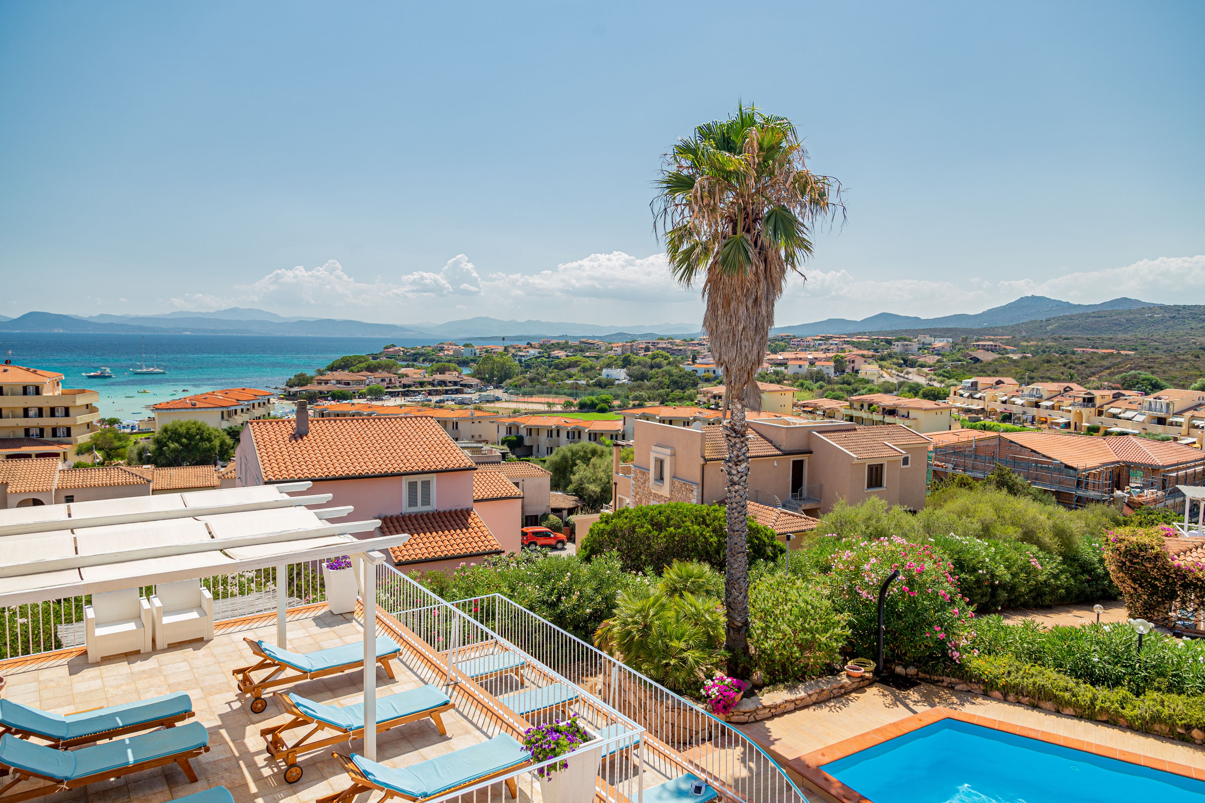 Una terraza en la azotea con sillones de descanso y piscina, con vistas a un pueblo costero con edificios y una palmera.