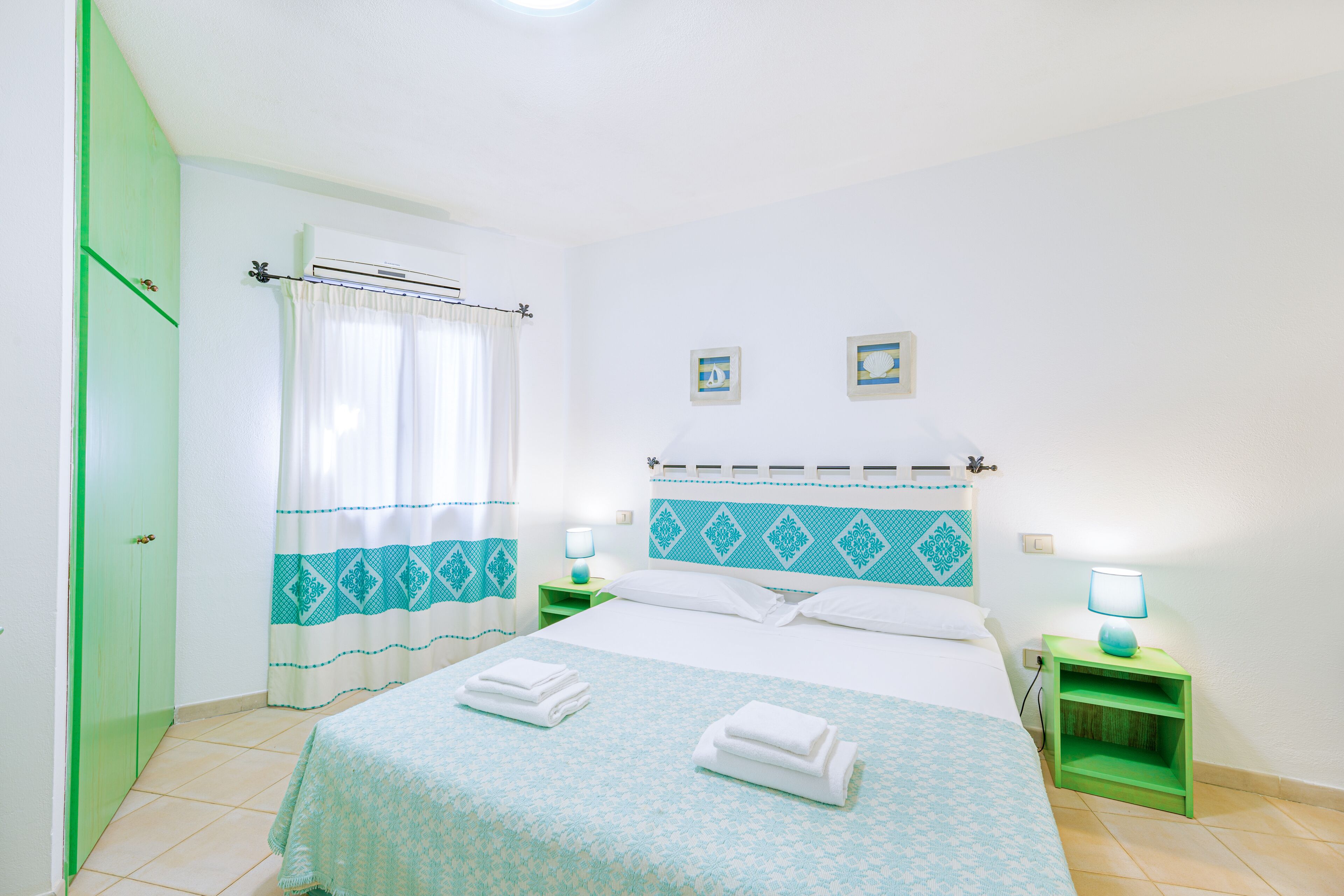 A neatly made bed with a patterned bedspread, two bedside tables with lamps, a green wardrobe, and a window with curtains.