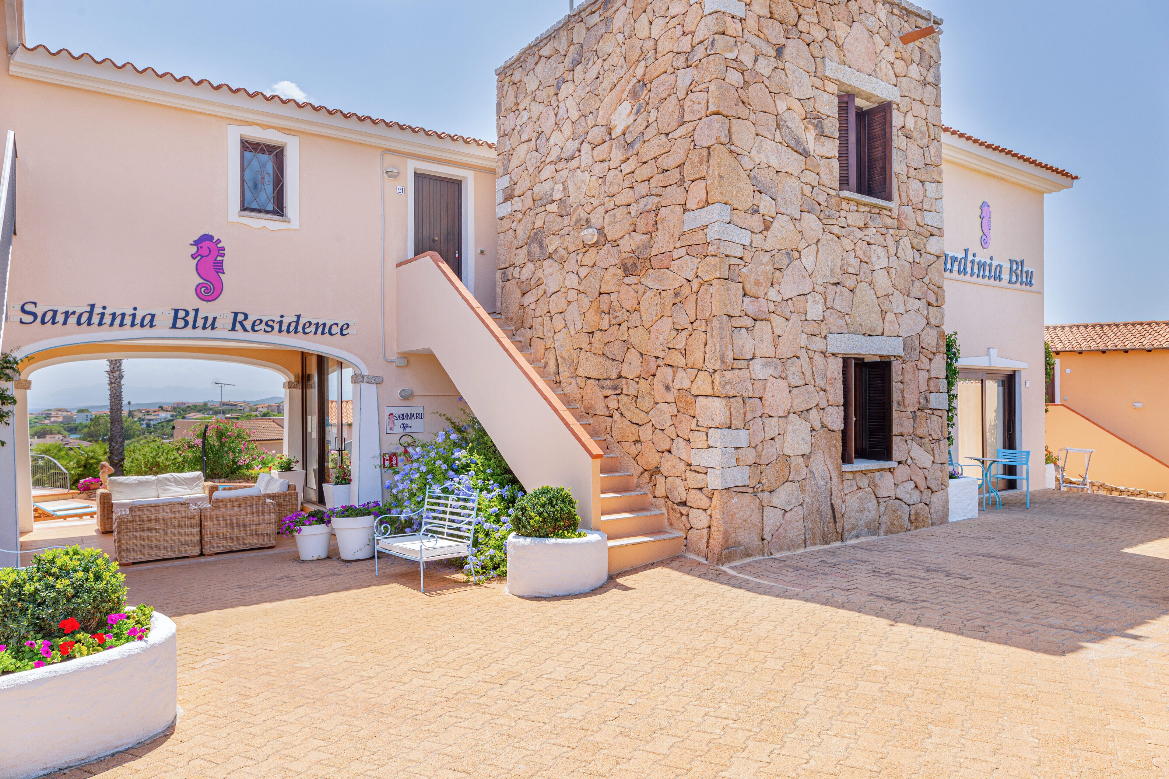 A building with a stone facade, a staircase leading to the entrance, outdoor seating, and potted plants.