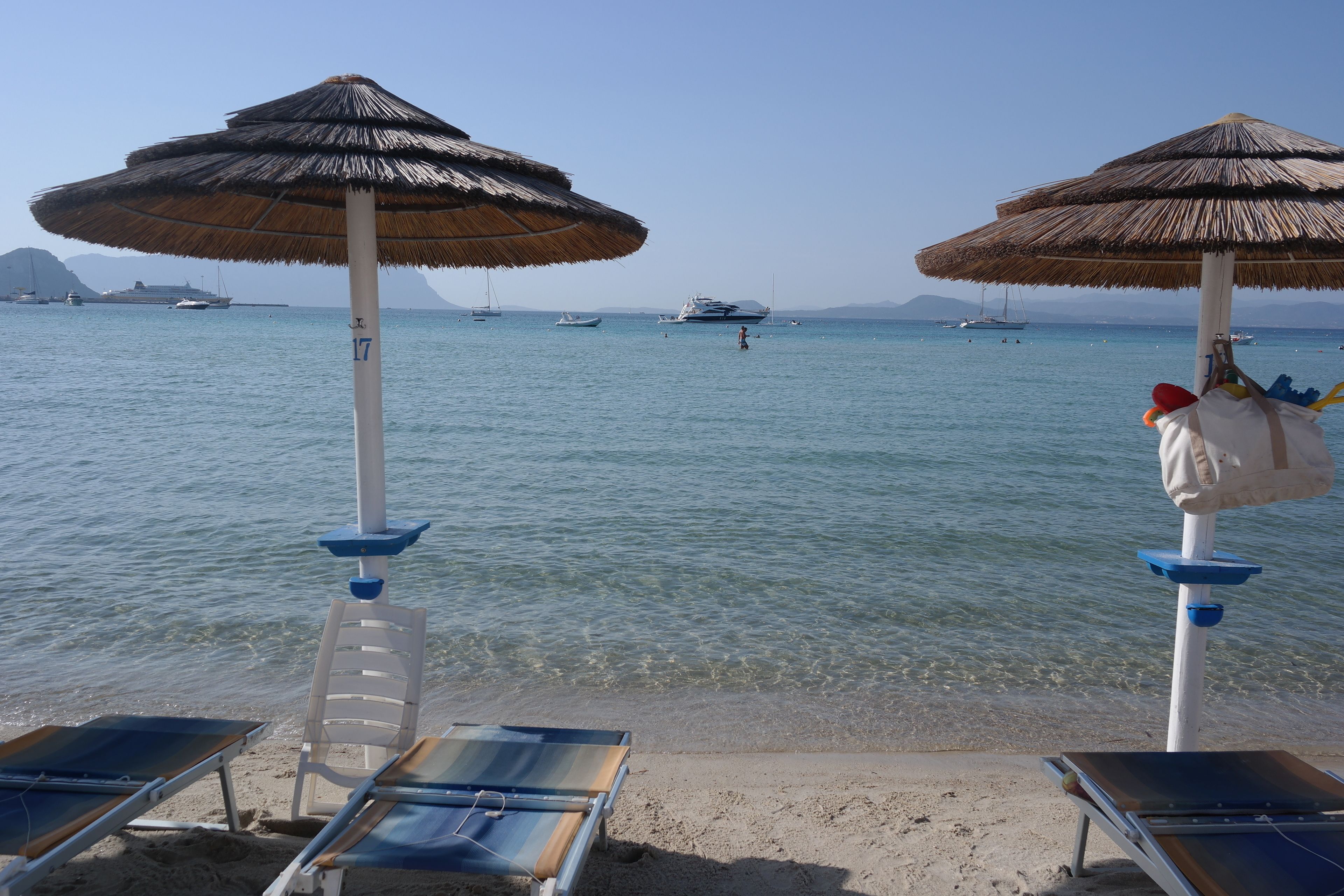A beach with two umbrellas, a row of sun loungers, and a clear view of the sea with boats in the distance.