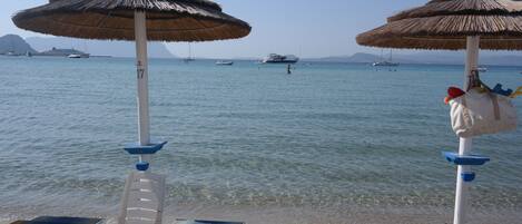 A beach with two umbrellas, a row of sun loungers, and a clear view of the sea with boats in the distance.