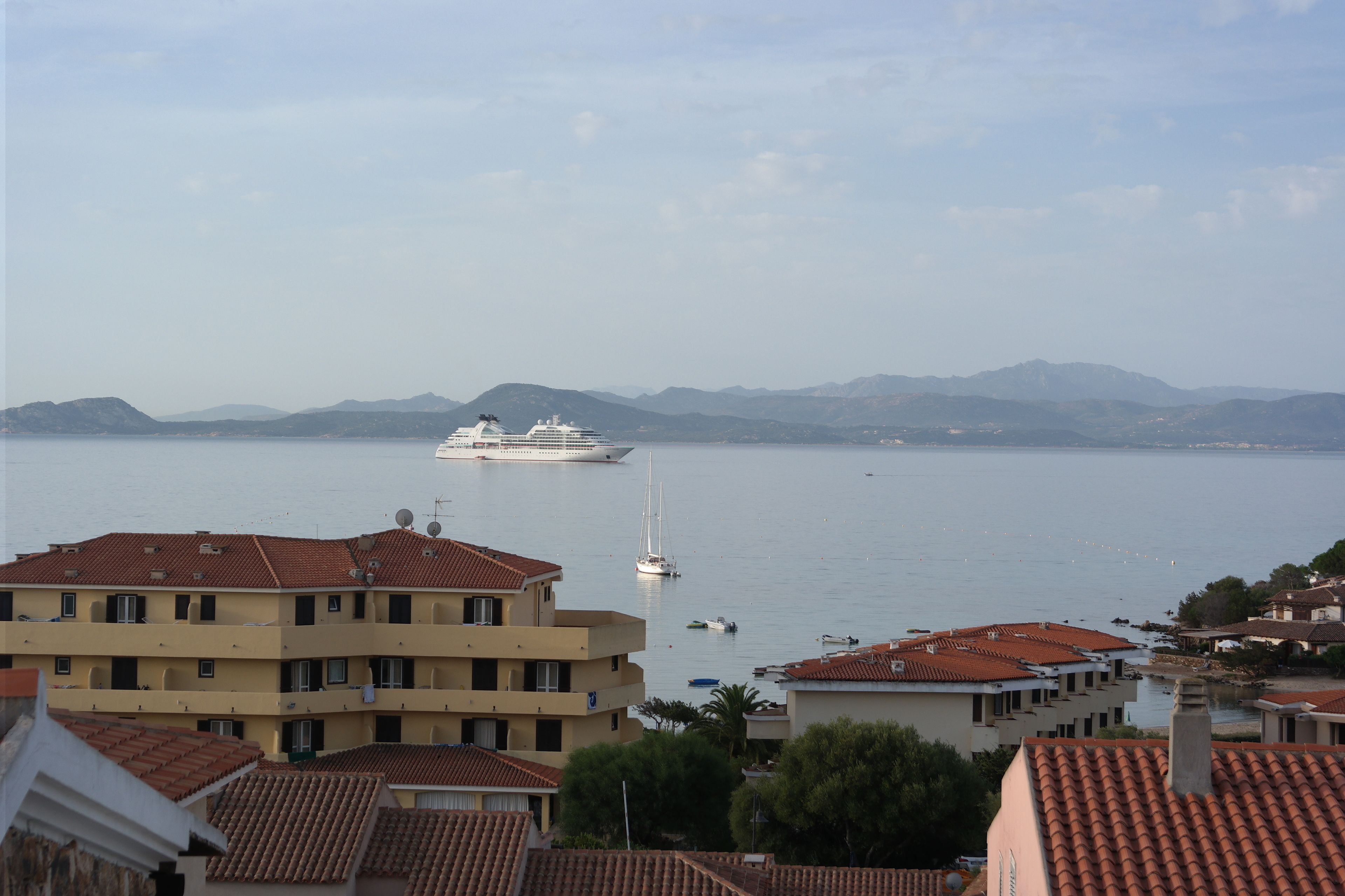A coastal town with buildings featuring red-tiled roofs, a marina with boats, and distant mountains.