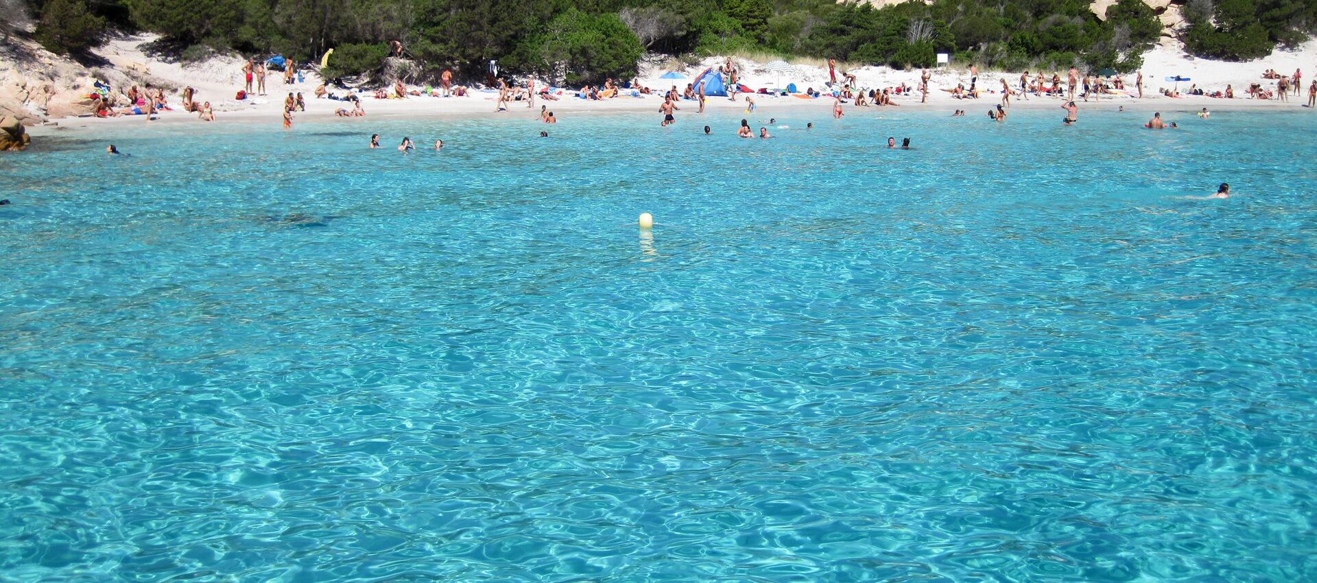 En strand med kristallklart blått vatten där folk badar och solar.