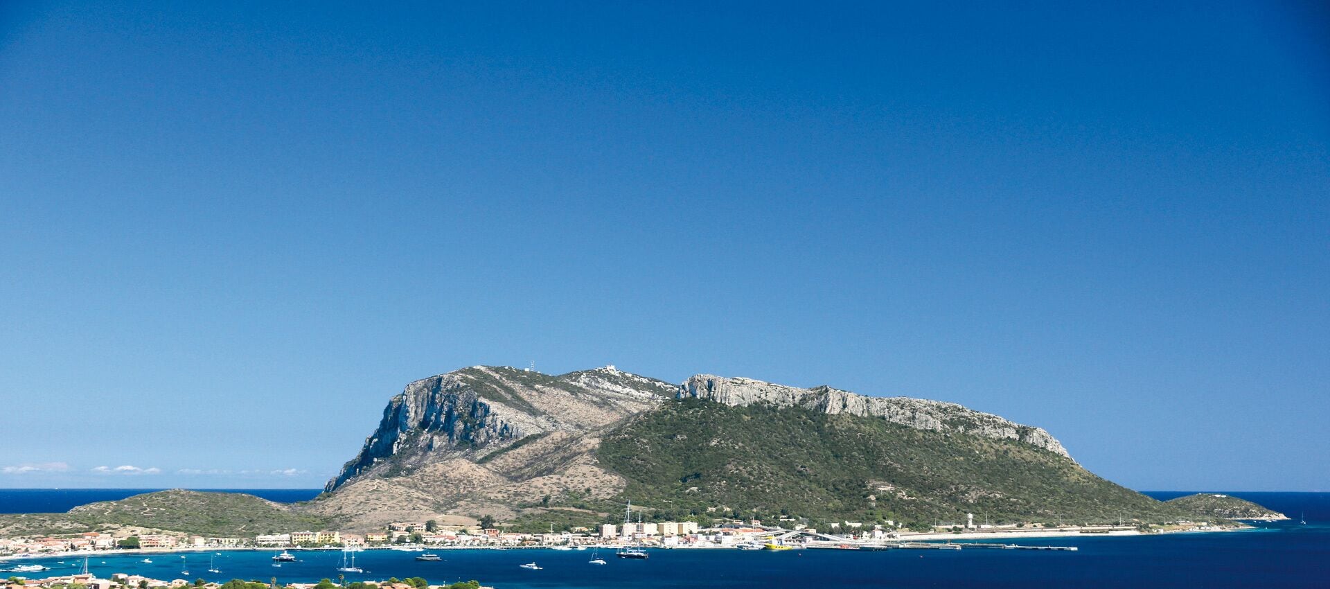 Une ville côtière caractérisée par une colline rocheuse imposante, une mer d’un bleu limpide et un port de plaisance où sont amarrés des bateaux.