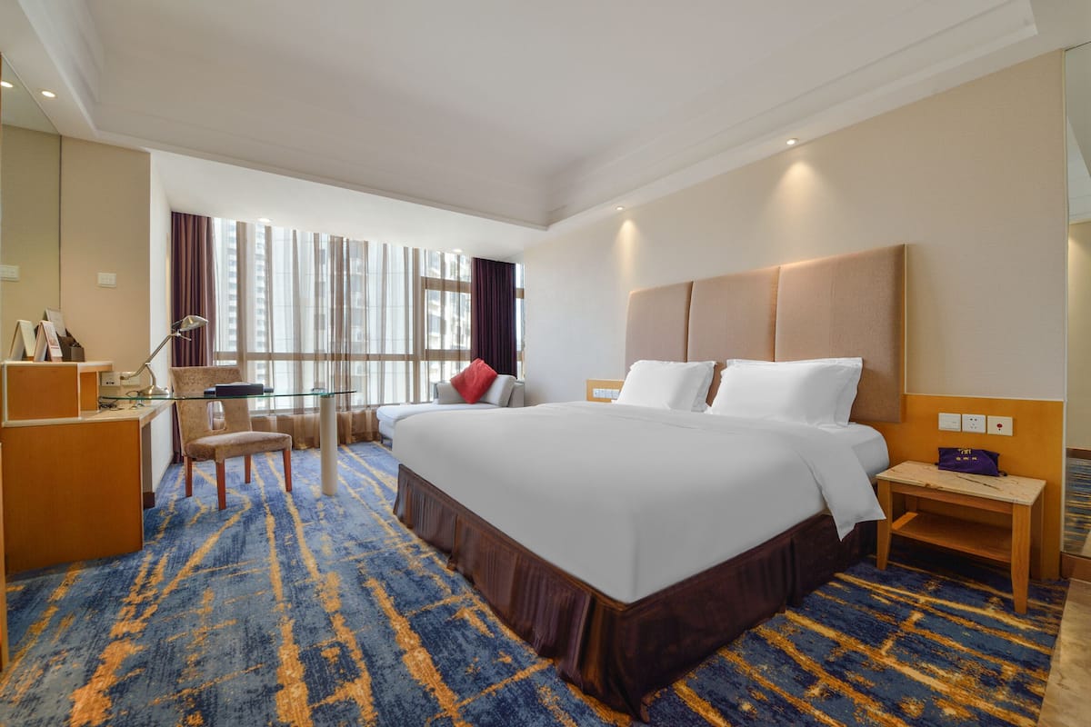 A hotel room with a large bed, a desk, and a chair. There is a window with curtains and a view of buildings outside.