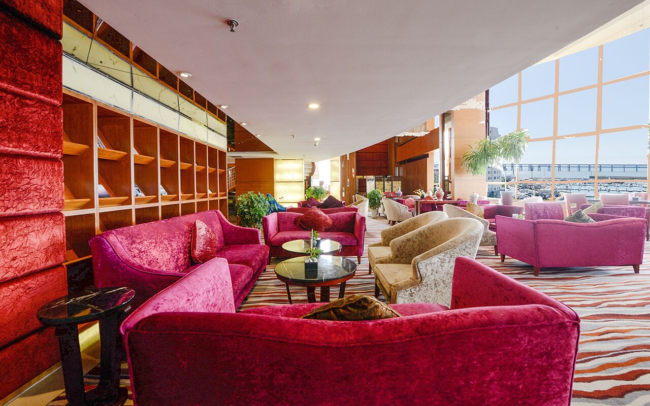 Eine großzügige Lobby mit roten, gepolsterten Sofas, einem zentralen Couchtisch und einem großen Fenster mit Blick auf den Strand.