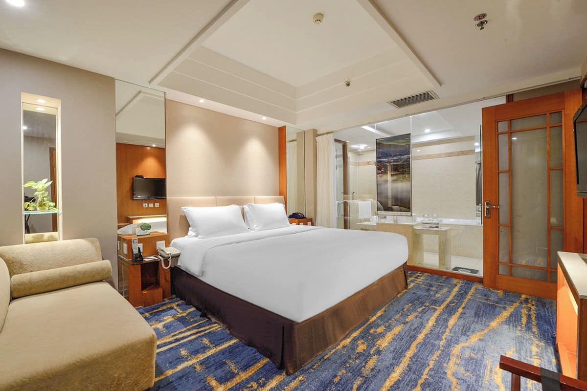 A hotel room with a large bed, a sofa, a desk with a telephone, a mirror, and a television.