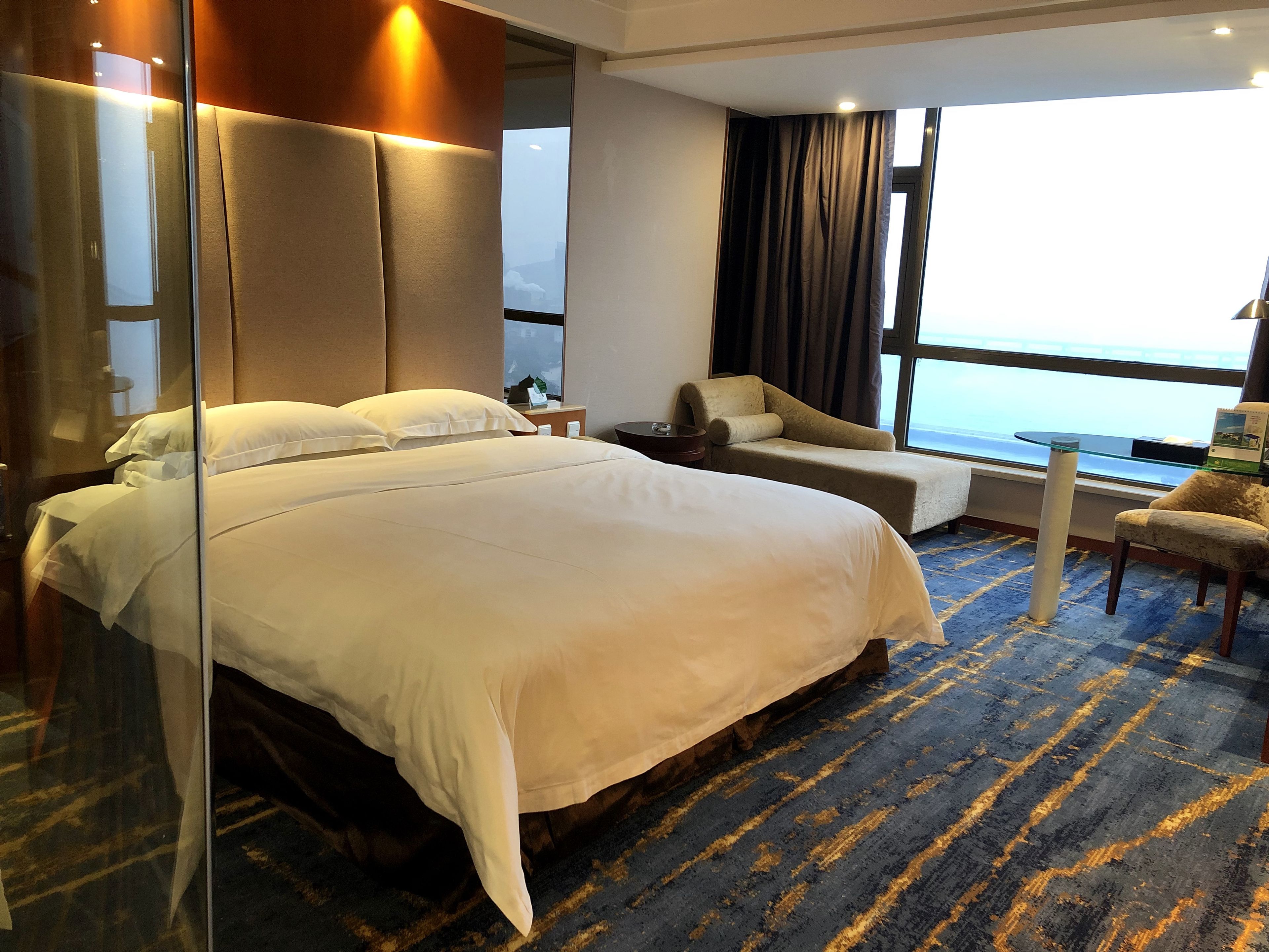 Una habitación de hotel con una cama grande, una zona de estar con una silla y una mesita, un ventanal con vista al mar y un separador de vidrio.