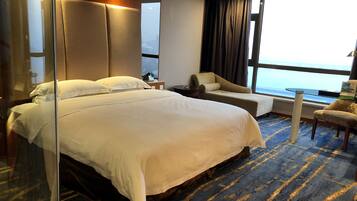 Una habitación de hotel con una cama grande, una zona de estar con una silla y una mesita, un ventanal con vista al mar y un separador de vidrio.