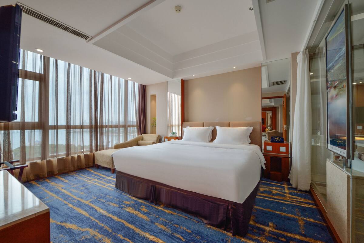 A hotel room with a large bed, a desk, and a view of the outdoors through large windows.
