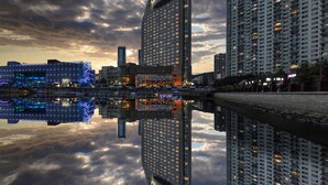 Un edificio alto con fachada curva que se refleja en el agua, rodeado de otros edificios esbeltos y un cielo nublado.