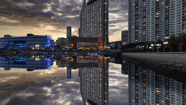 A high-rise building with a curved facade reflecting in a body of water, surrounded by other tall buildings and a cloudy sky. - Bayshore Hotel Dalian (Dalian)