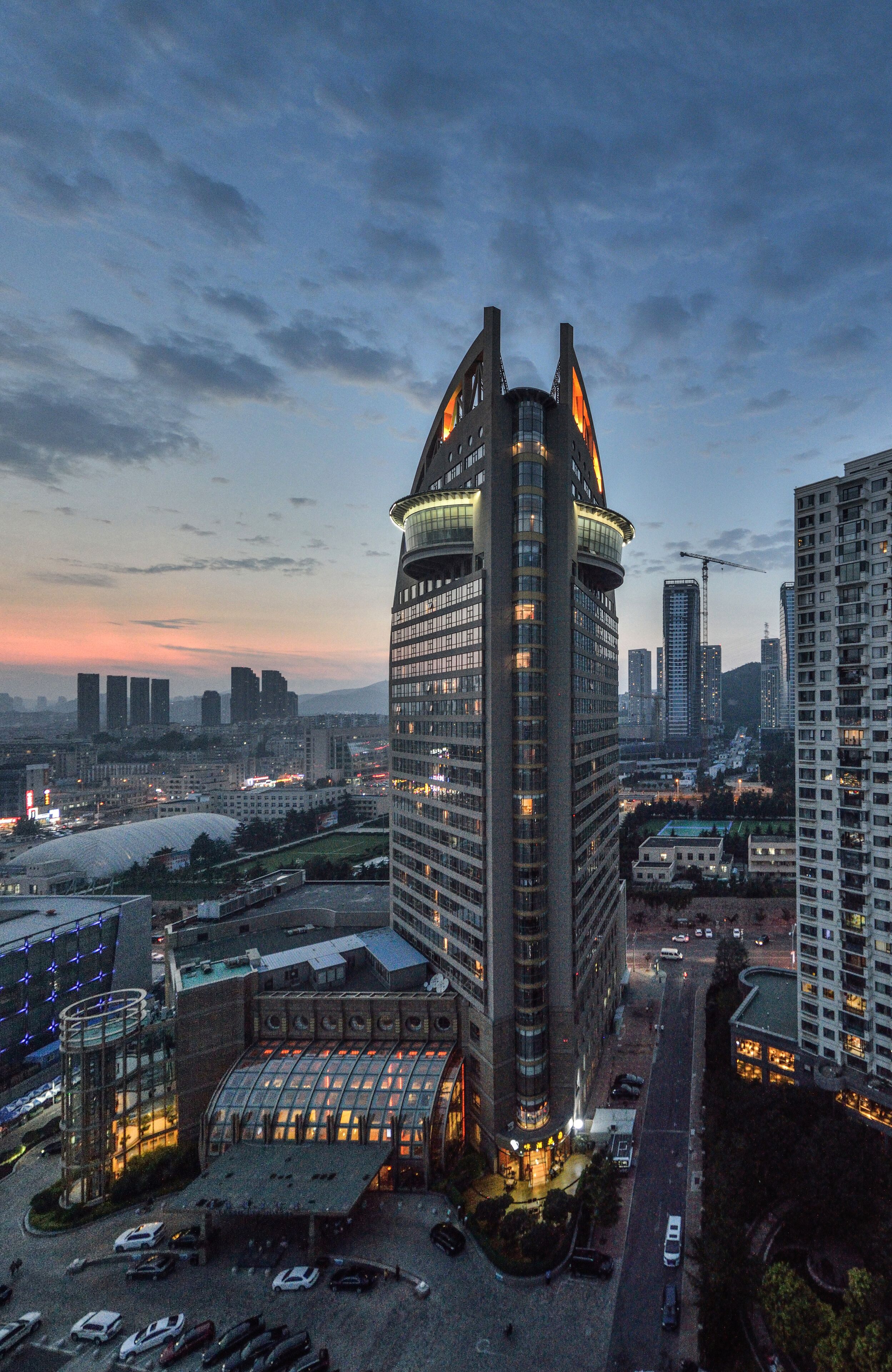 A high-rise building with a distinctive curved top and multiple windows, surrounded by other tall buildings and a parking area.
