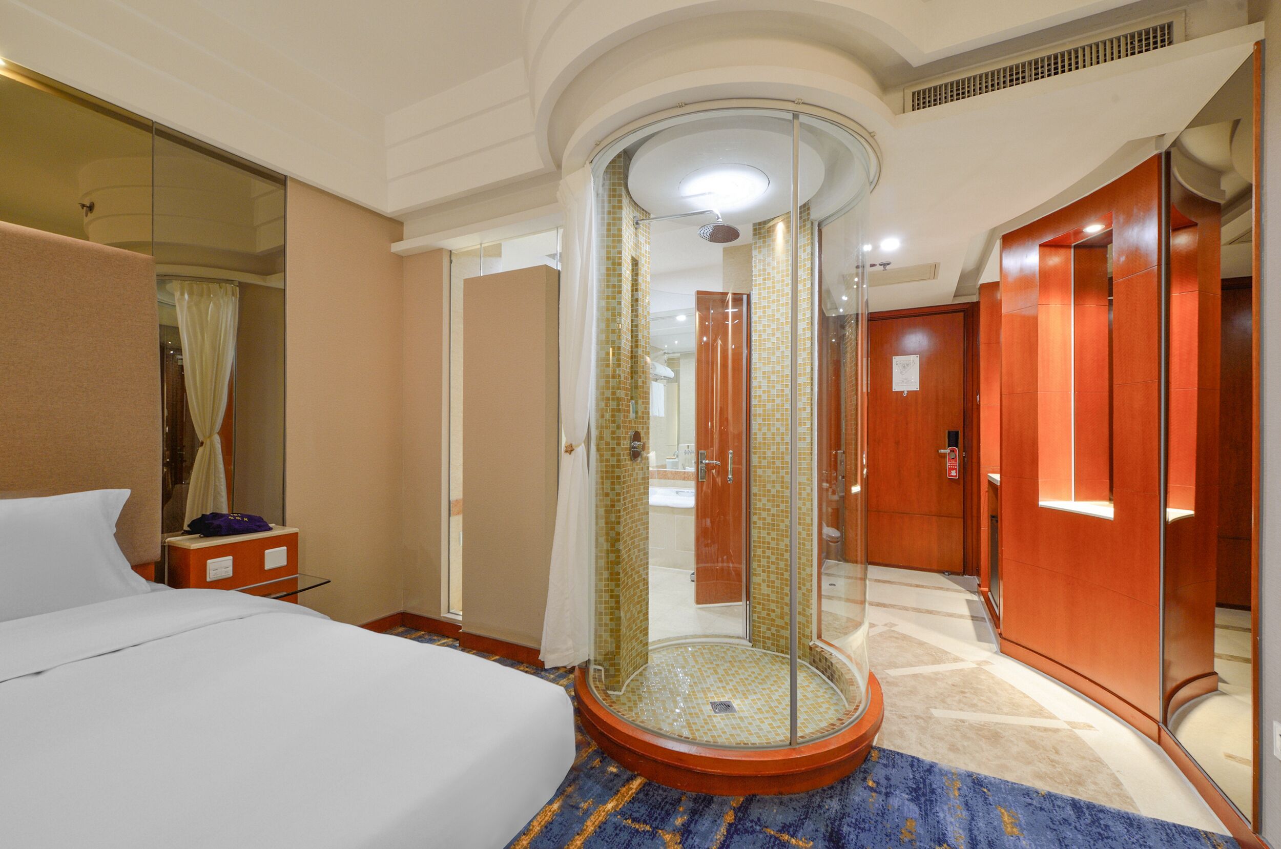 Una habitación de hotel con cama, ducha envuelta en vidrio, armario de madera y baño con espejo y lavabo.