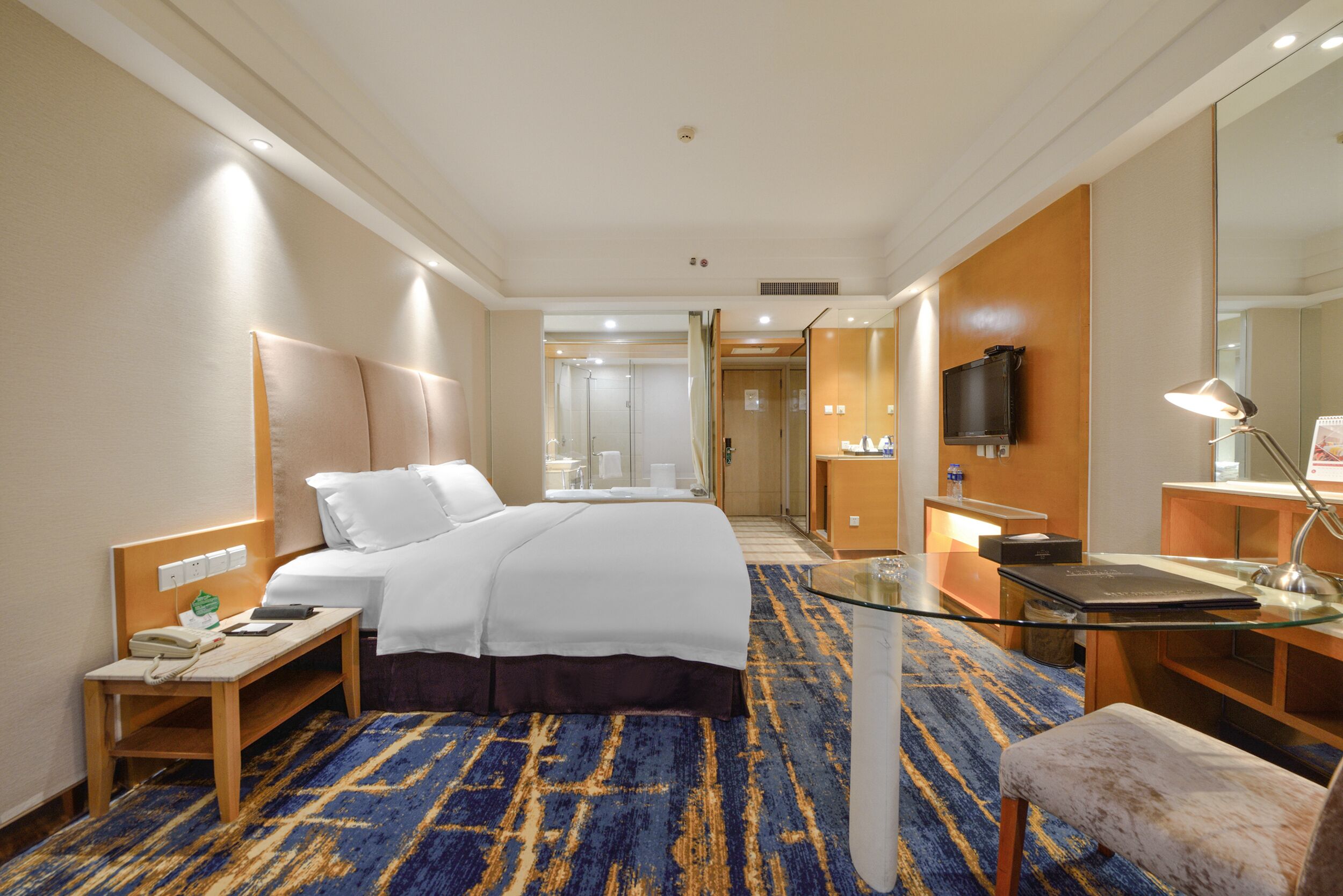 A hotel room with a large bed, a desk with a lamp, a TV, and a bathroom visible through an open door.