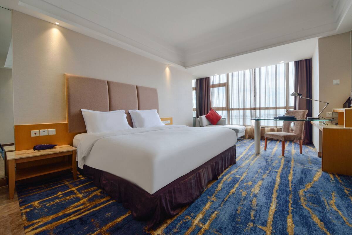 A hotel room with a large bed, a desk, a chair, and a window with curtains.