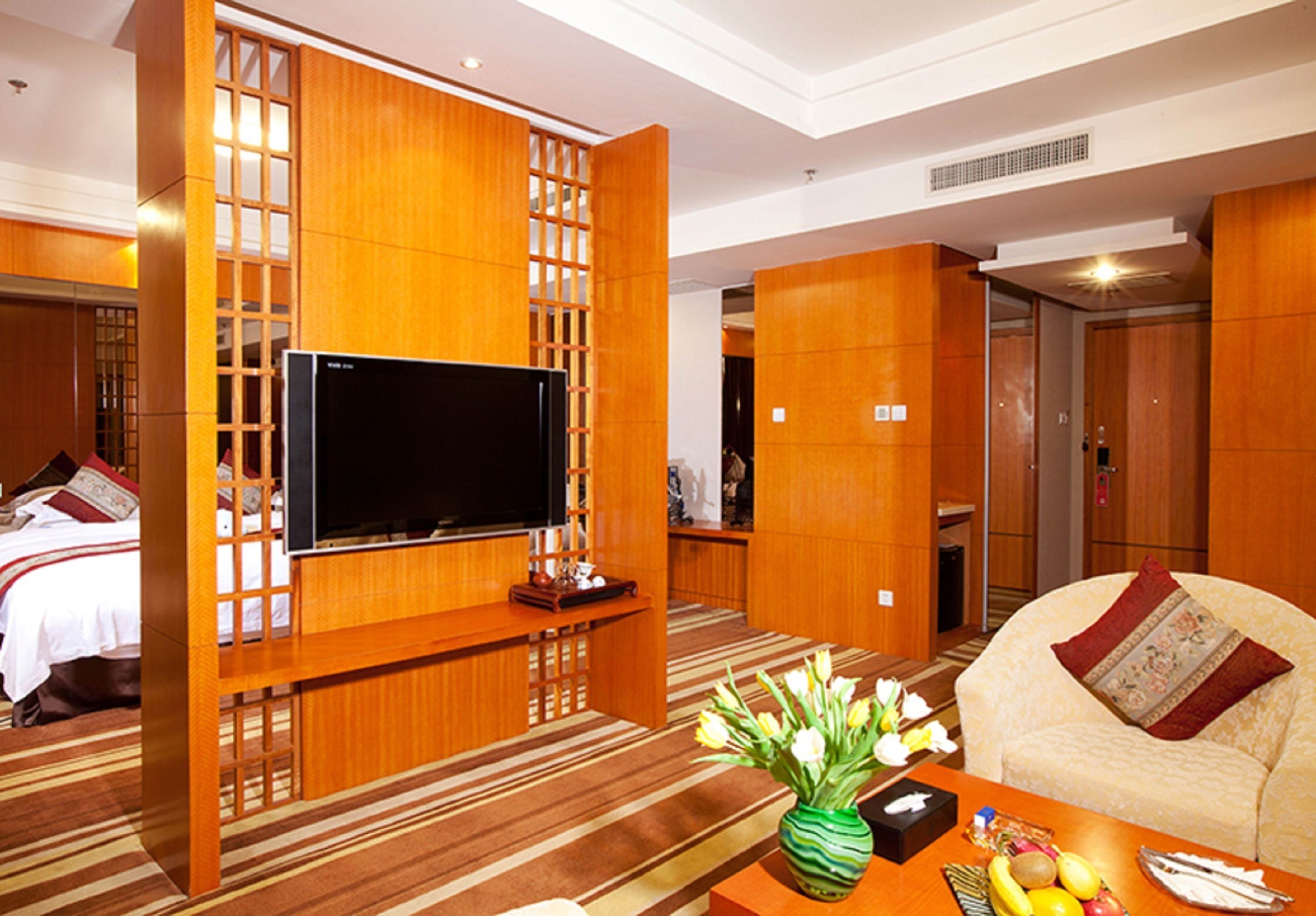 A hotel room with a TV mounted on a wooden partition, a sofa, a coffee table with fruits and a vase of flowers, and a bed with a red pillow.