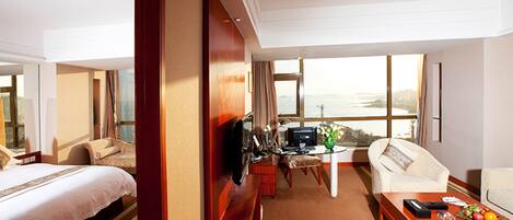 A hotel room with a bed, a desk with a computer, a chair, and a view of the sea.