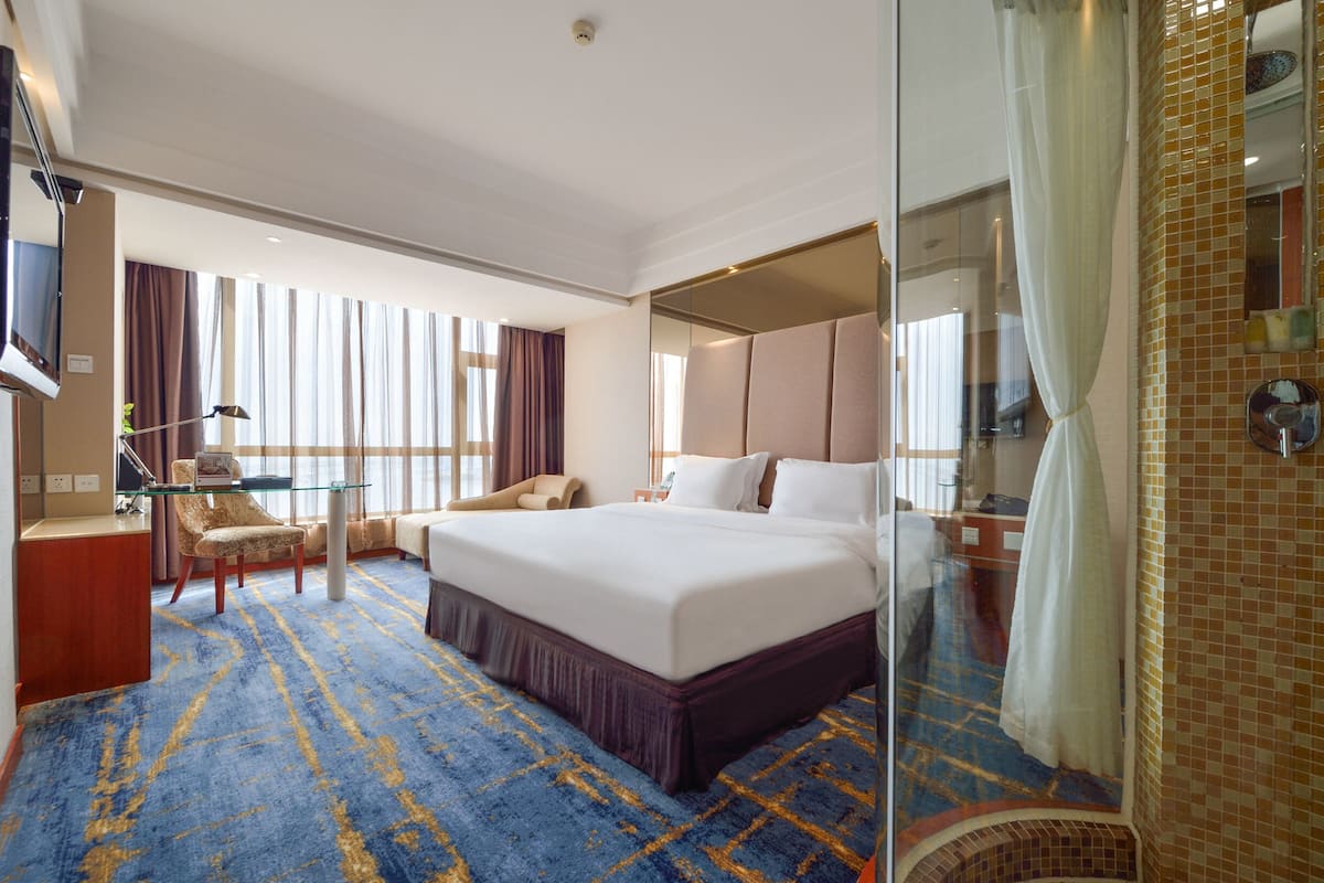 A hotel room with a large bed, a desk with a chair, a TV, and a bathroom with a shower and a mirror.