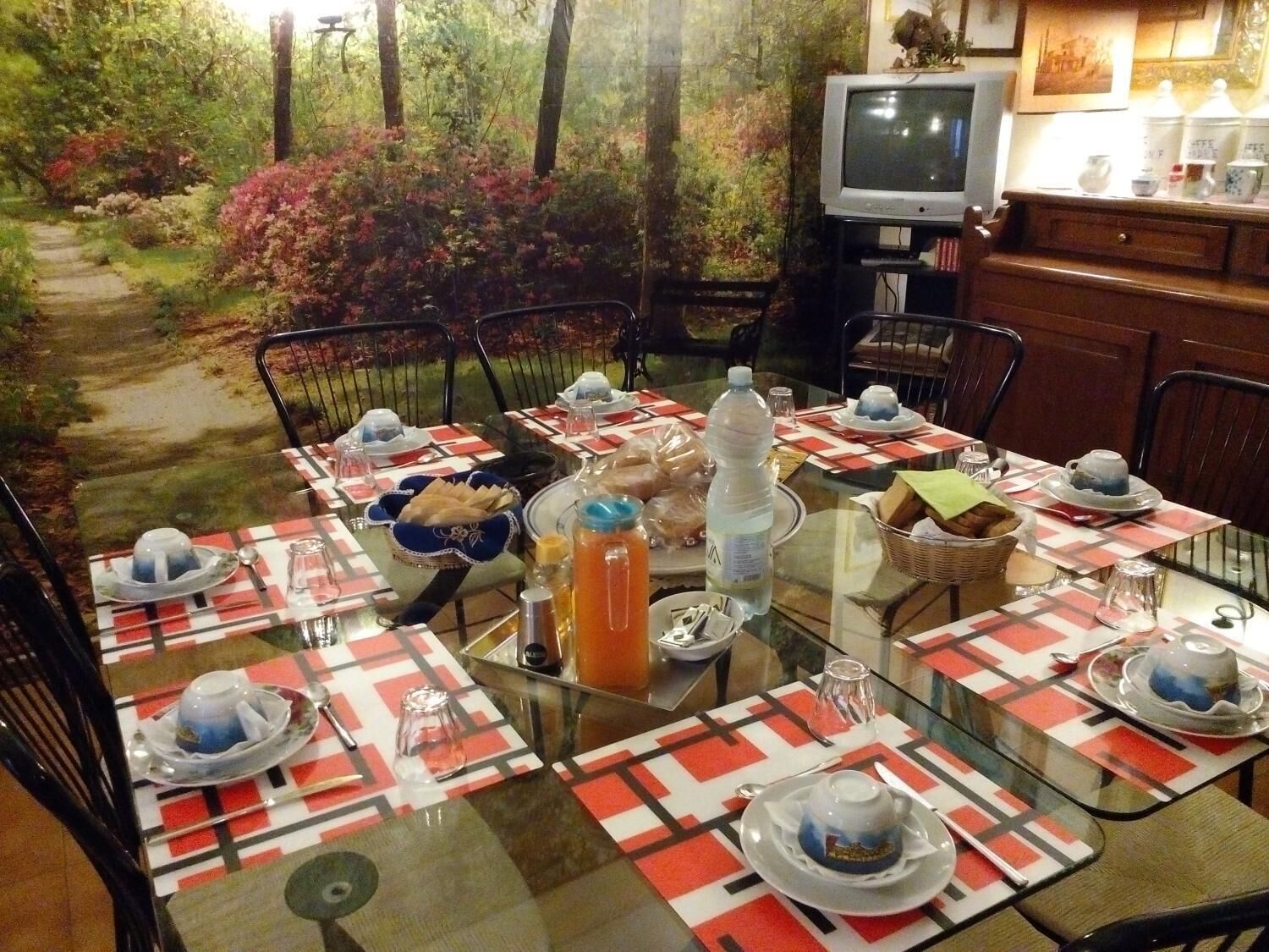 A dining area with a glass table set for four, surrounded by chairs, with a television and kitchen area in the background.