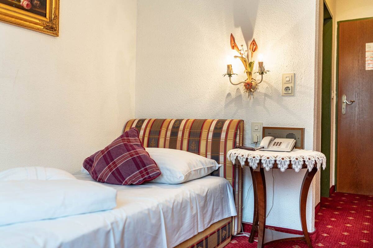 Un lit simple avec une couverture à carreaux et un oreiller rouge, une table de chevet avec un téléphone et un luminaire fixé au mur.
