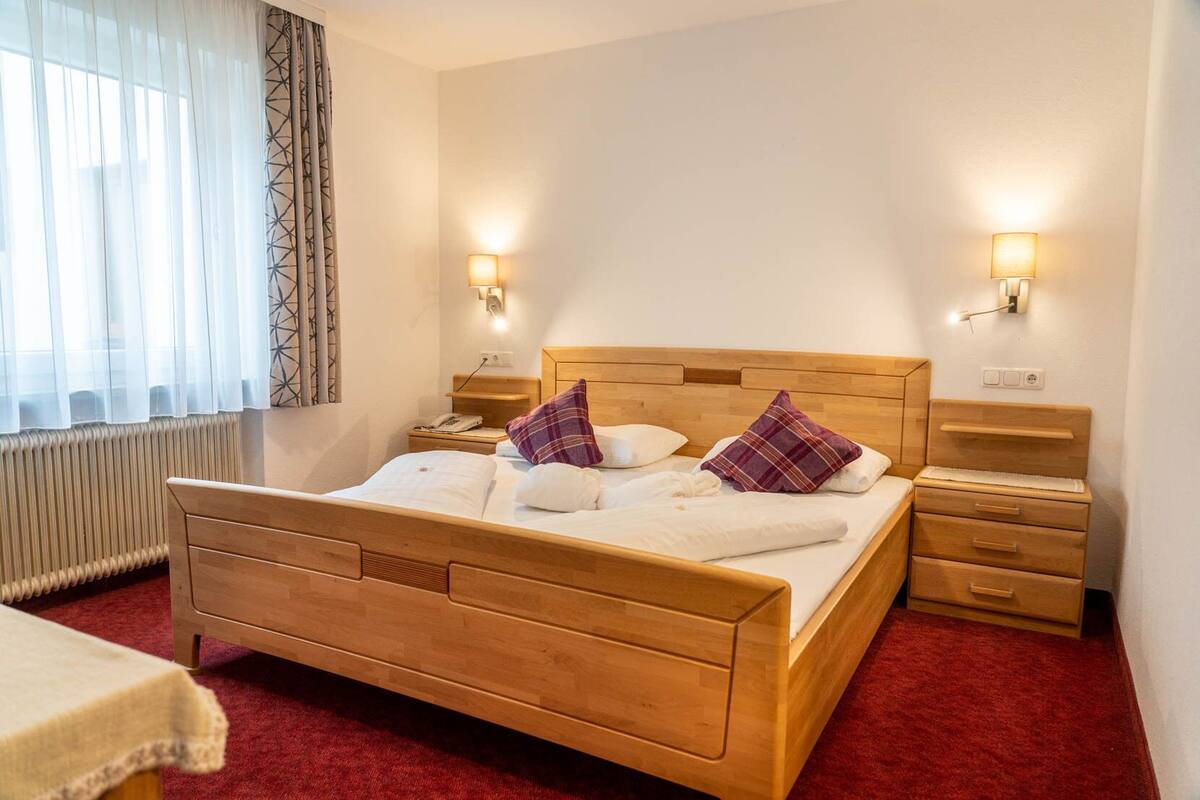 Une chambre d’hôtel avec un lit en bois, deux tables de chevet, une petite table, une moquette rouge, un radiateur blanc et une fenêtre avec des rideaux légers.