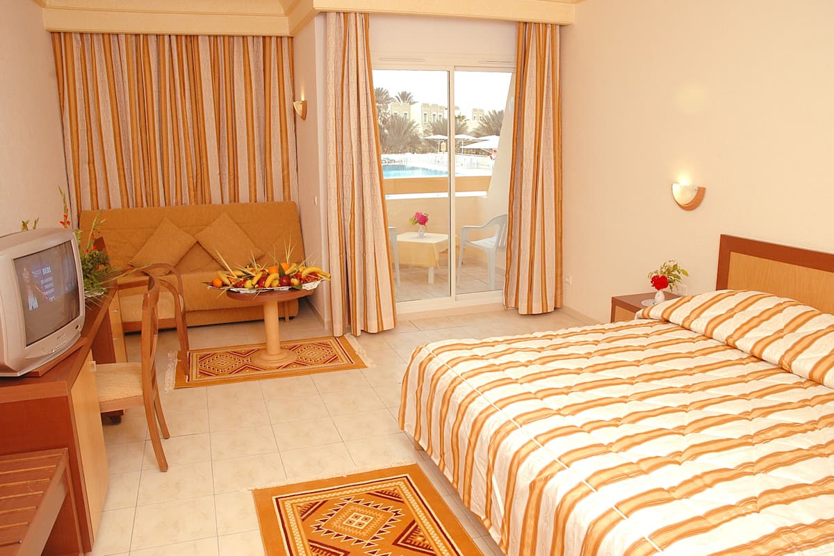 A hotel room with a bed, a sofa, a TV, a small table with fruit, and a balcony with a pool view.