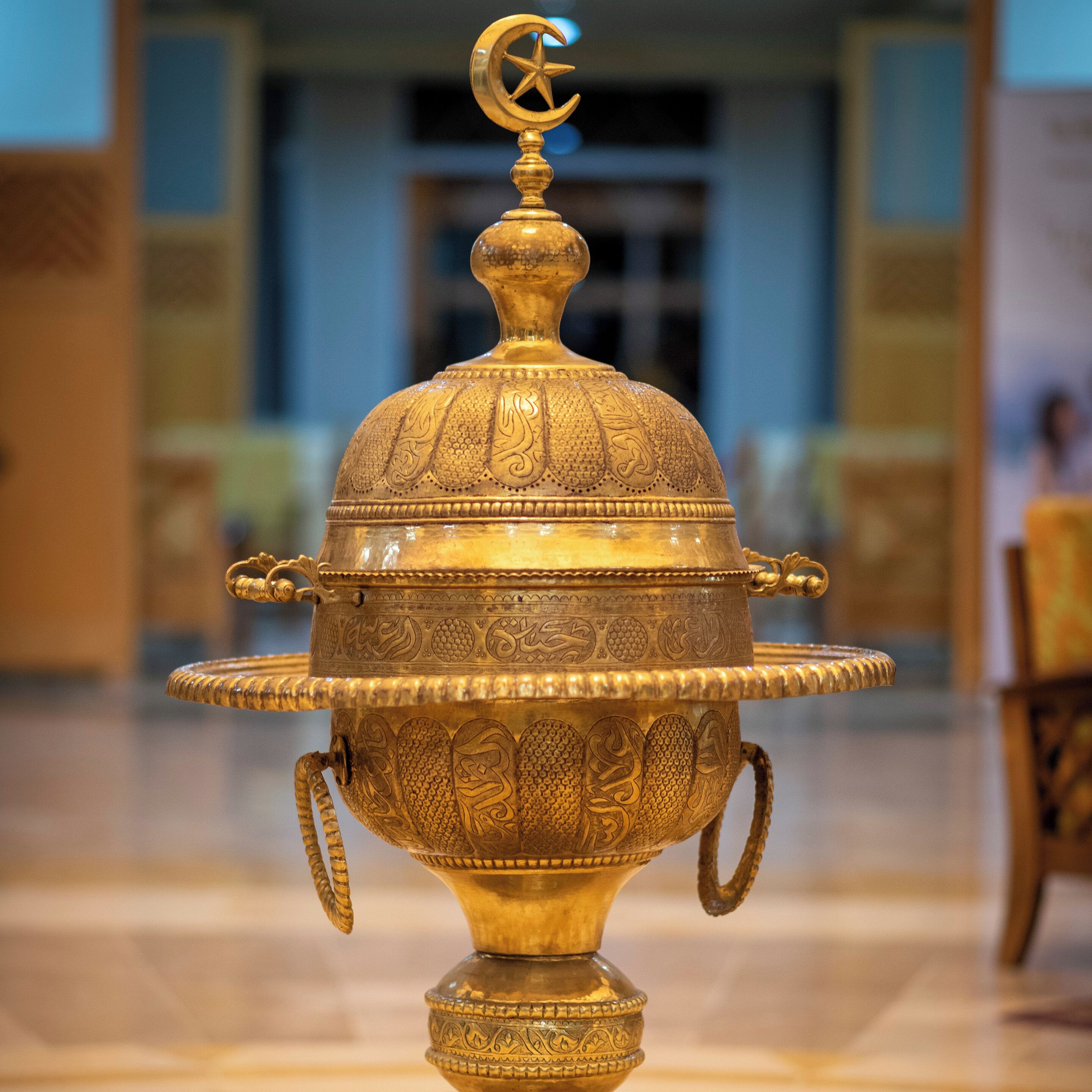 A brass artifact with intricate designs and a crescent moon symbol on top, placed on a tiled floor.