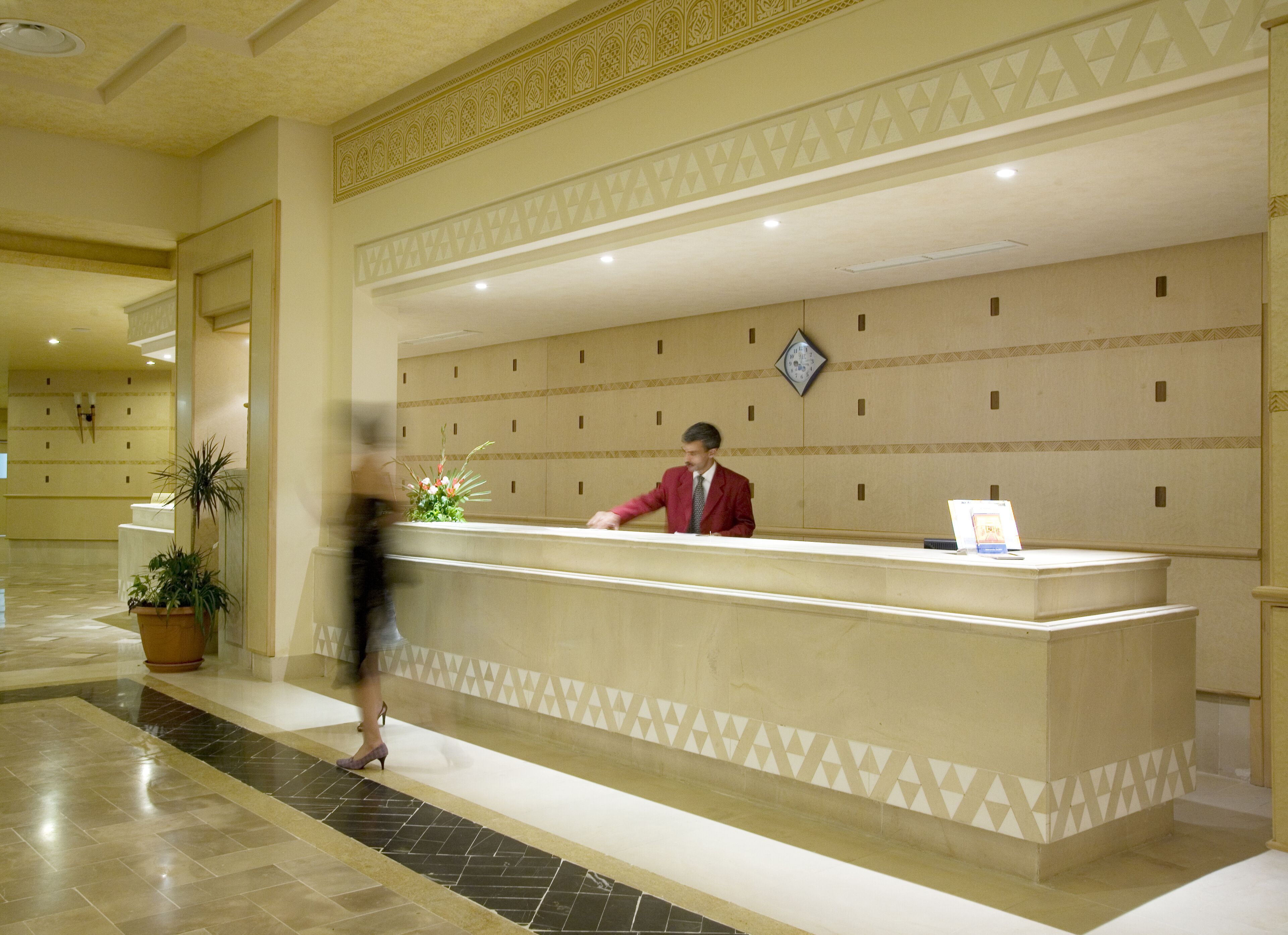 A hotel reception desk with a man in a red suit standing behind it. There is a clock on the wall above the desk and a potted plant to the left.