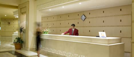 A hotel reception desk with a man in a red suit standing behind it. There is a clock on the wall above the desk and a potted plant to the left.