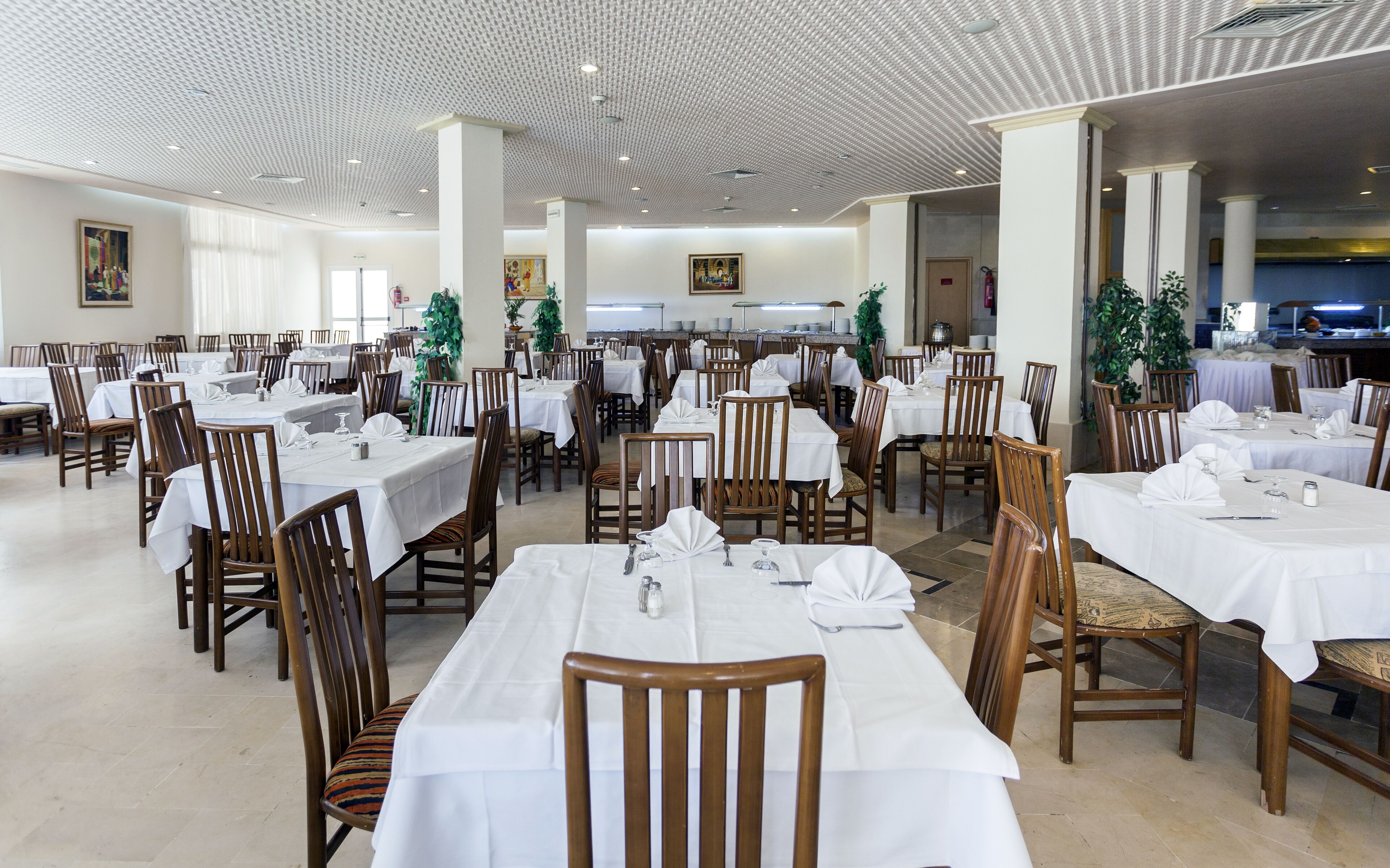 Een ruime eethoek met meerdere tafels en stoelen, een hoog plafond met een gedetailleerd patroon, en een keuken in de achtergrond.