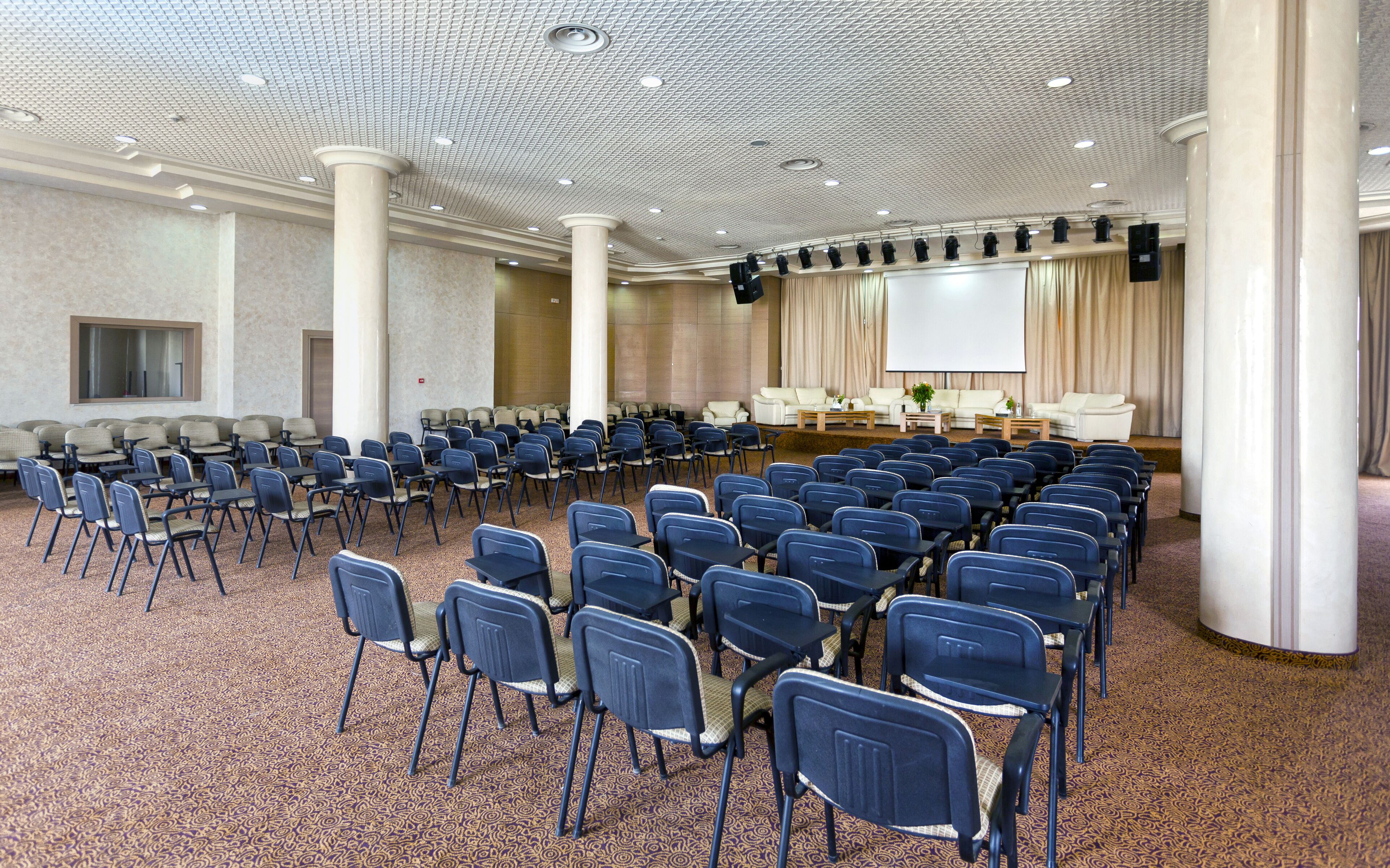 A conference room with rows of chairs, a stage with a projector, and a large screen.