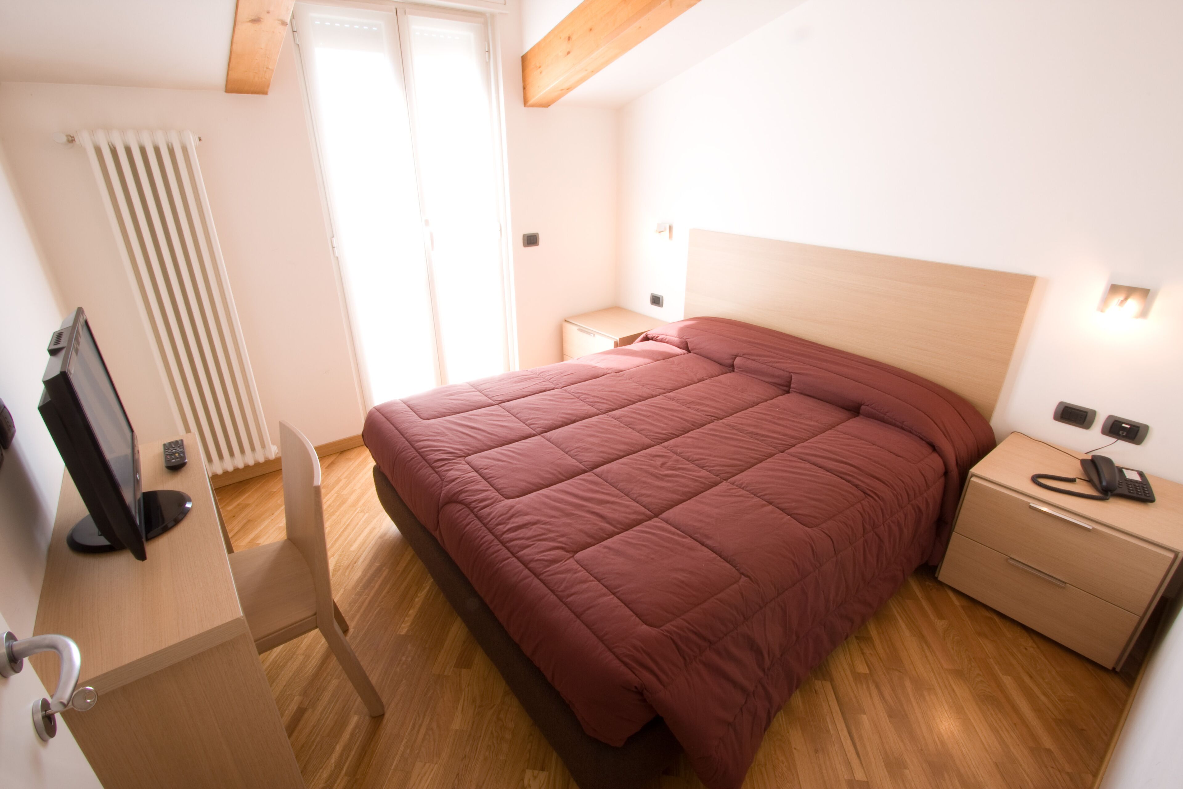 A bedroom with a bed, a nightstand, a desk with a computer, a radiator, and a window.