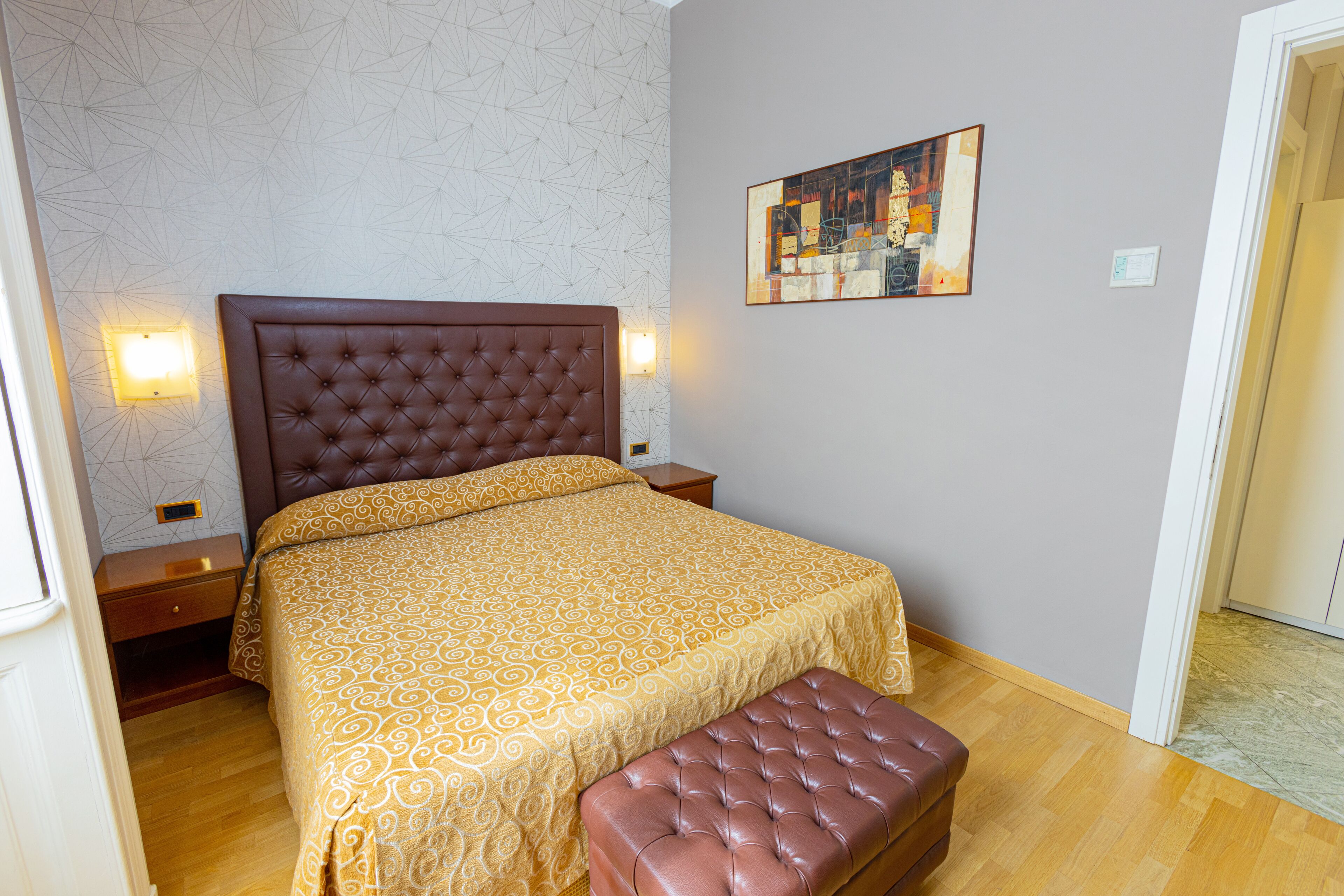 A bedroom with a large bed, a headboard, a bedside table, a nightstand, a wall-mounted light, and a painting.