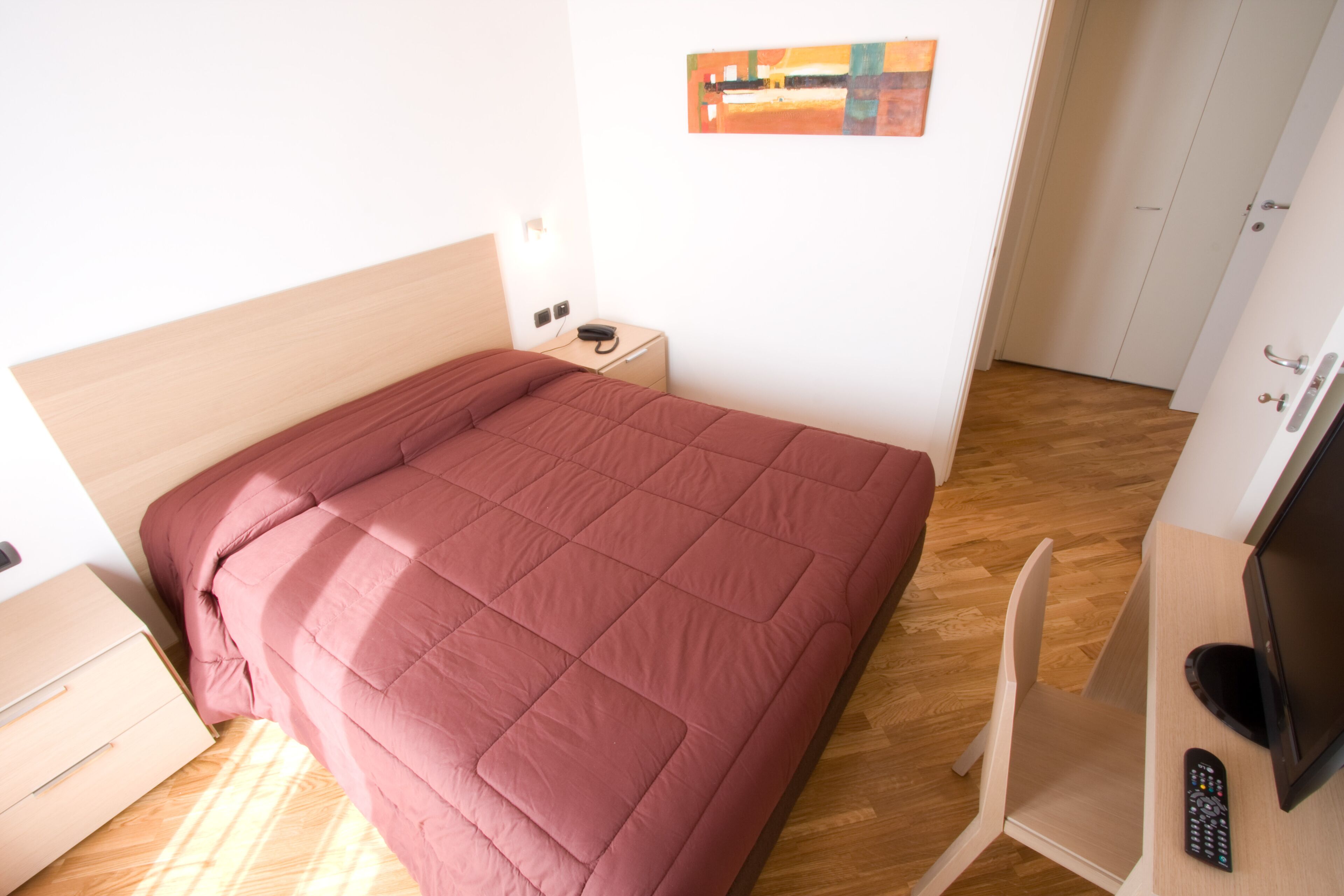 Una cama bien hecha con cabecera de madera, una mesita de noche con lámpara, un mueble para televisor con control remoto y piso de madera.