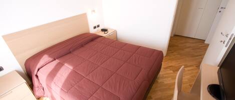 Una cama bien hecha con cabecera de madera, una mesita de noche con lámpara, un mueble para televisor con control remoto y piso de madera.