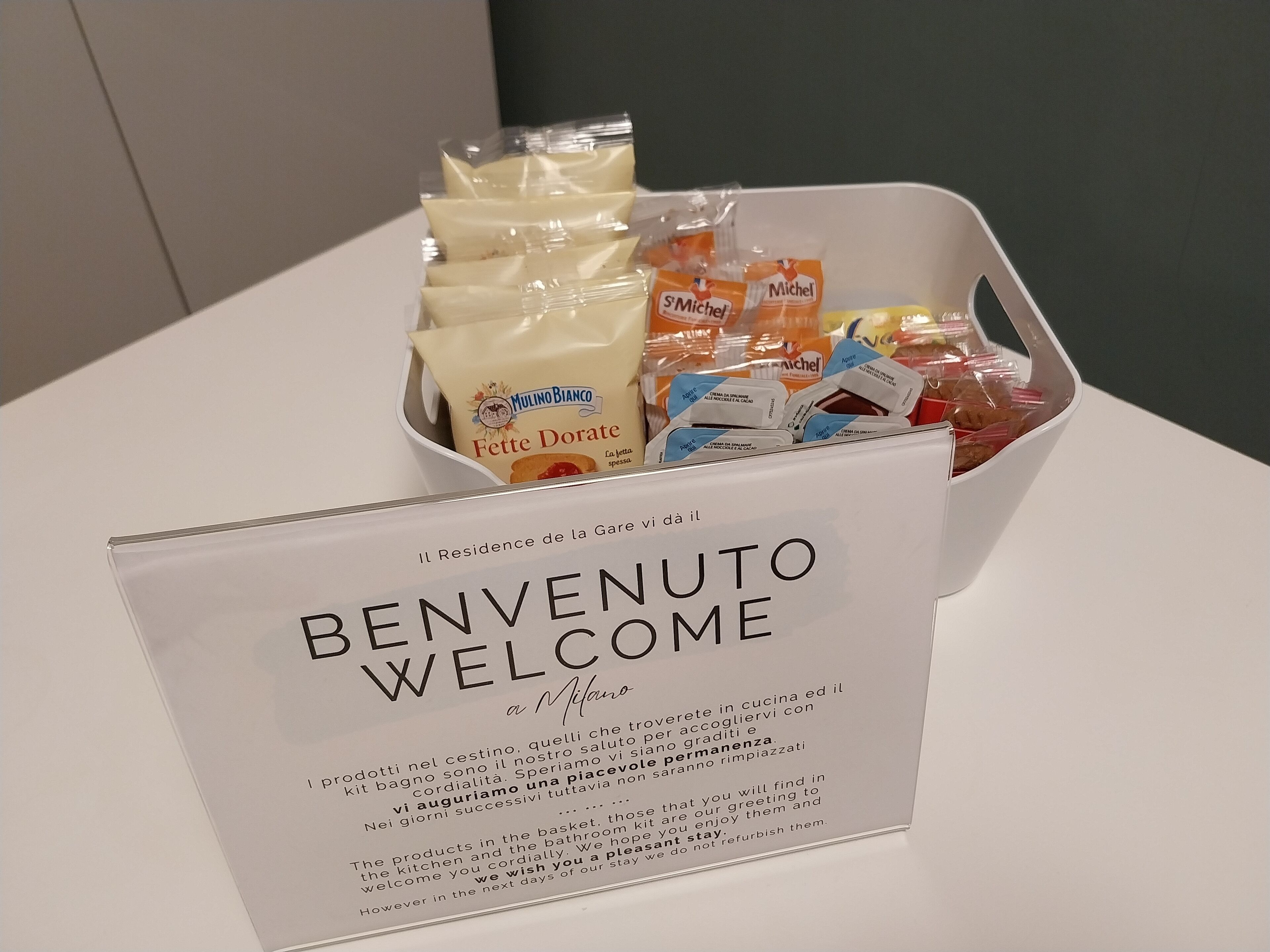 Een welkomstbord met een mand vol levensmiddelen, waaronder 'Mulino Bianco Fette Dorate' en 'Michel'.