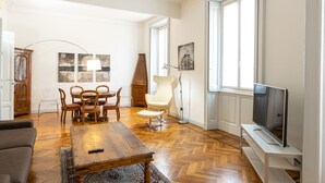 Una sala de estar con piso de madera, una mesa de comedor con sillas, un sofá, un televisor sobre un mueble bajo y obras de arte en las paredes.