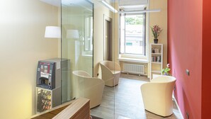 Una habitación de hotel moderna con un separador de vidrio, una cafetera, dos sillas, una estantería y una ventana con vistas al exterior.