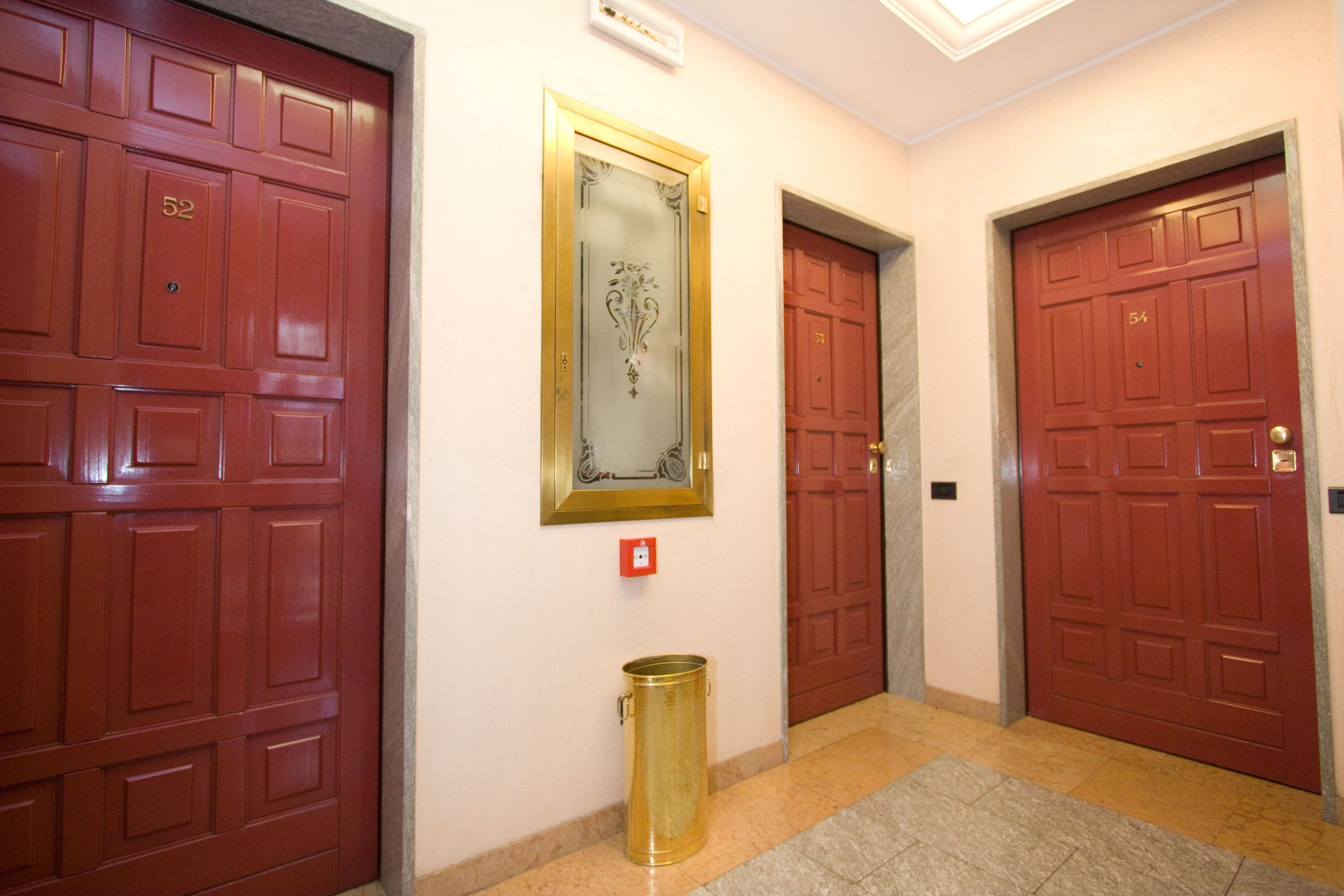Un couloir avec trois portes rouges, chacune portant un numéro. Une poubelle dorée est placée près de la porte du milieu.