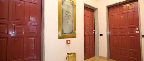 Un couloir avec trois portes rouges, chacune portant un numéro. Une poubelle dorée est placée près de la porte du milieu.