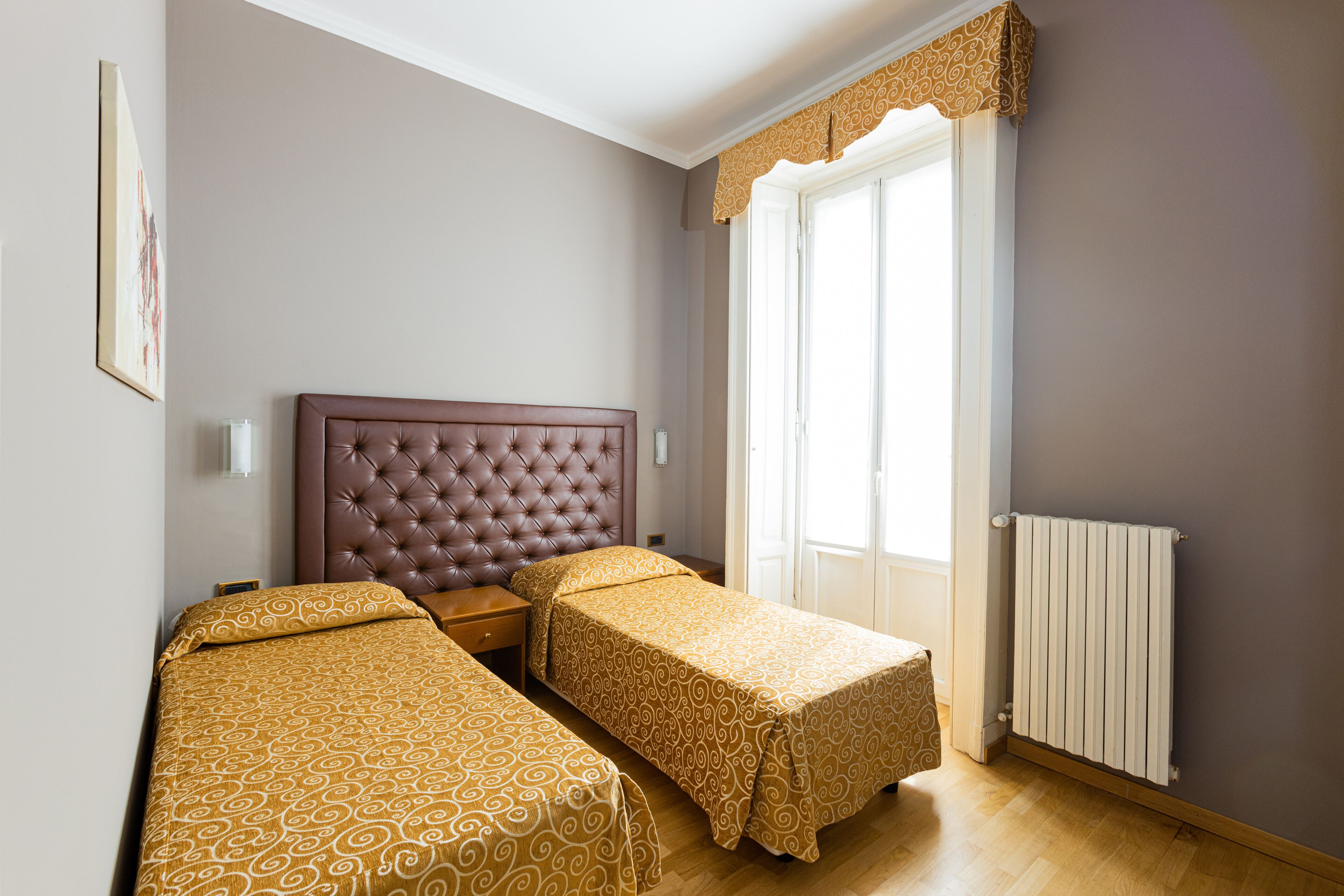 A bedroom with two single beds, a large window with a curtain, a radiator, and a framed picture on the wall.