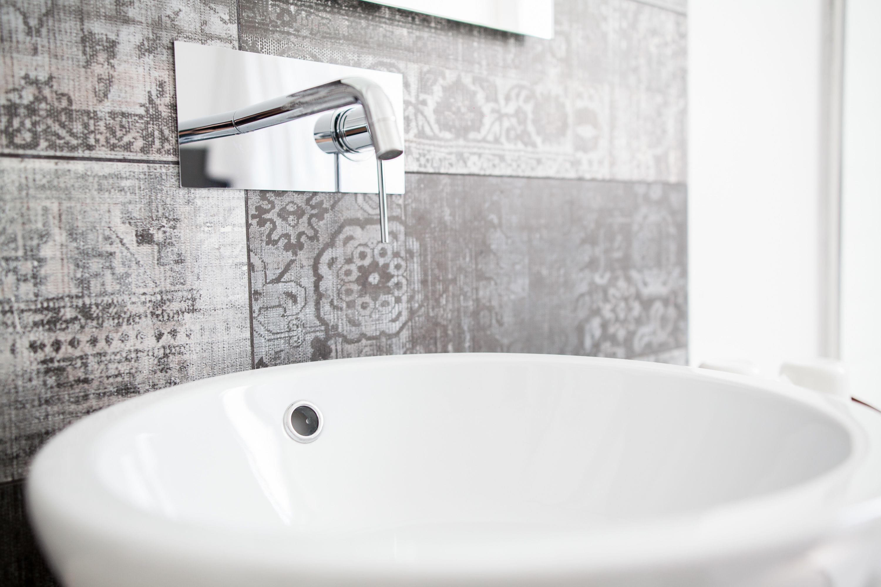 A bathroom with a white sink, a chrome faucet, and patterned tiles on the wall.