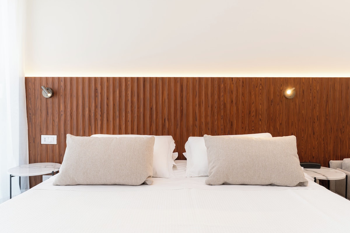 Una cama bien hecha con dos almohadas, un cabecero de madera y una mesita redonda a su lado.