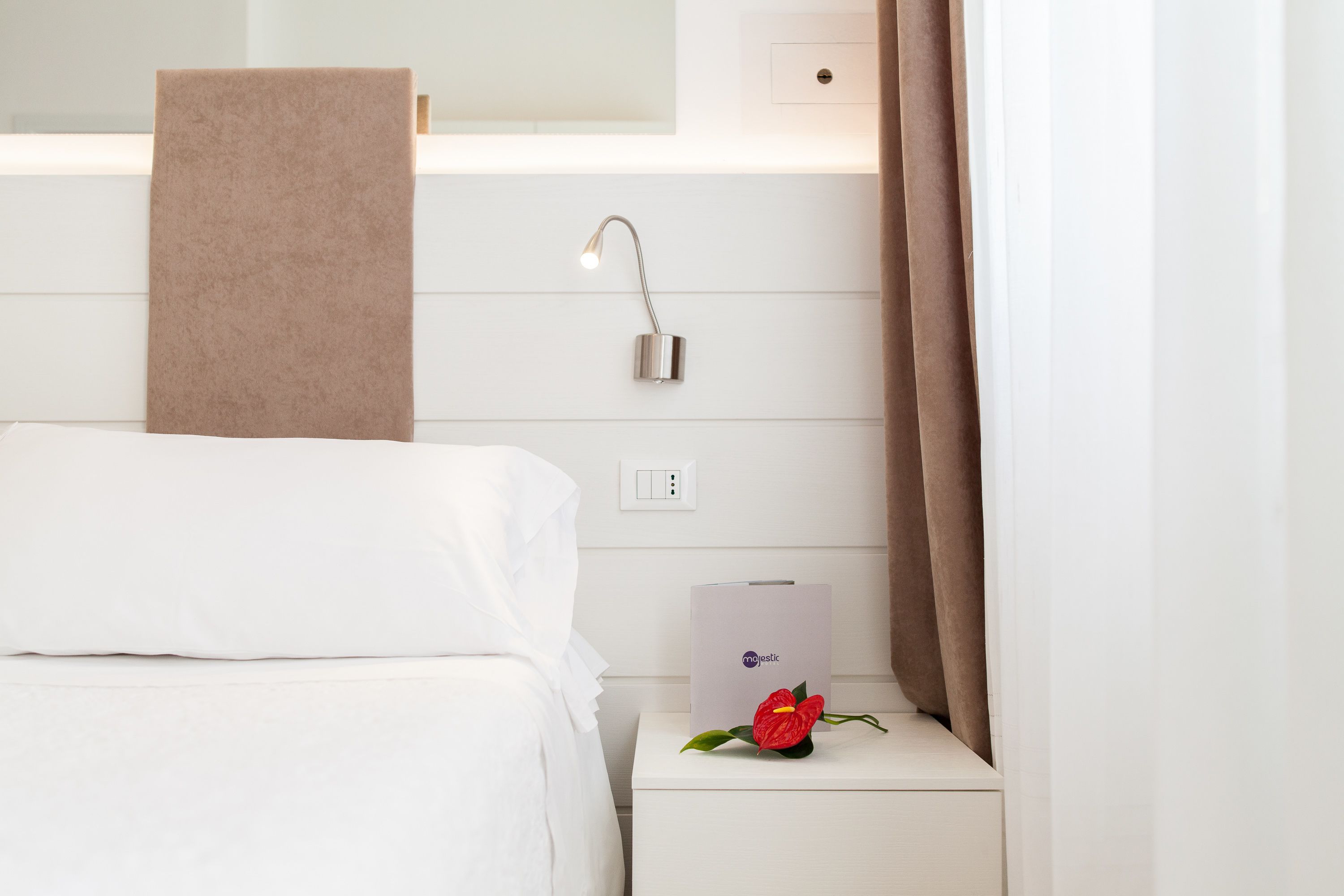 A hotel room with a neatly made bed, a bedside table with a lamp, a towel hanging on the wall, and a flower on the table.